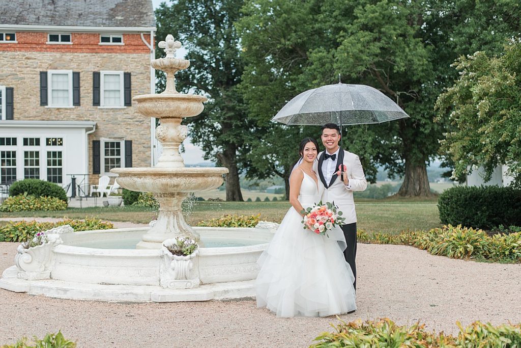 Bride and groom standing under a clear umbrella beside a stone fountain at Springfield Manor Winery.
