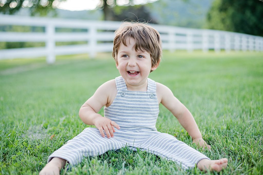 Smiling toddler boy in striped overalls sitting in a grassy field during a golden hour family photo session in Fluvanna County, Virginia. Captured by a Charlottesville family photographer known for relaxed, outdoor portraits.