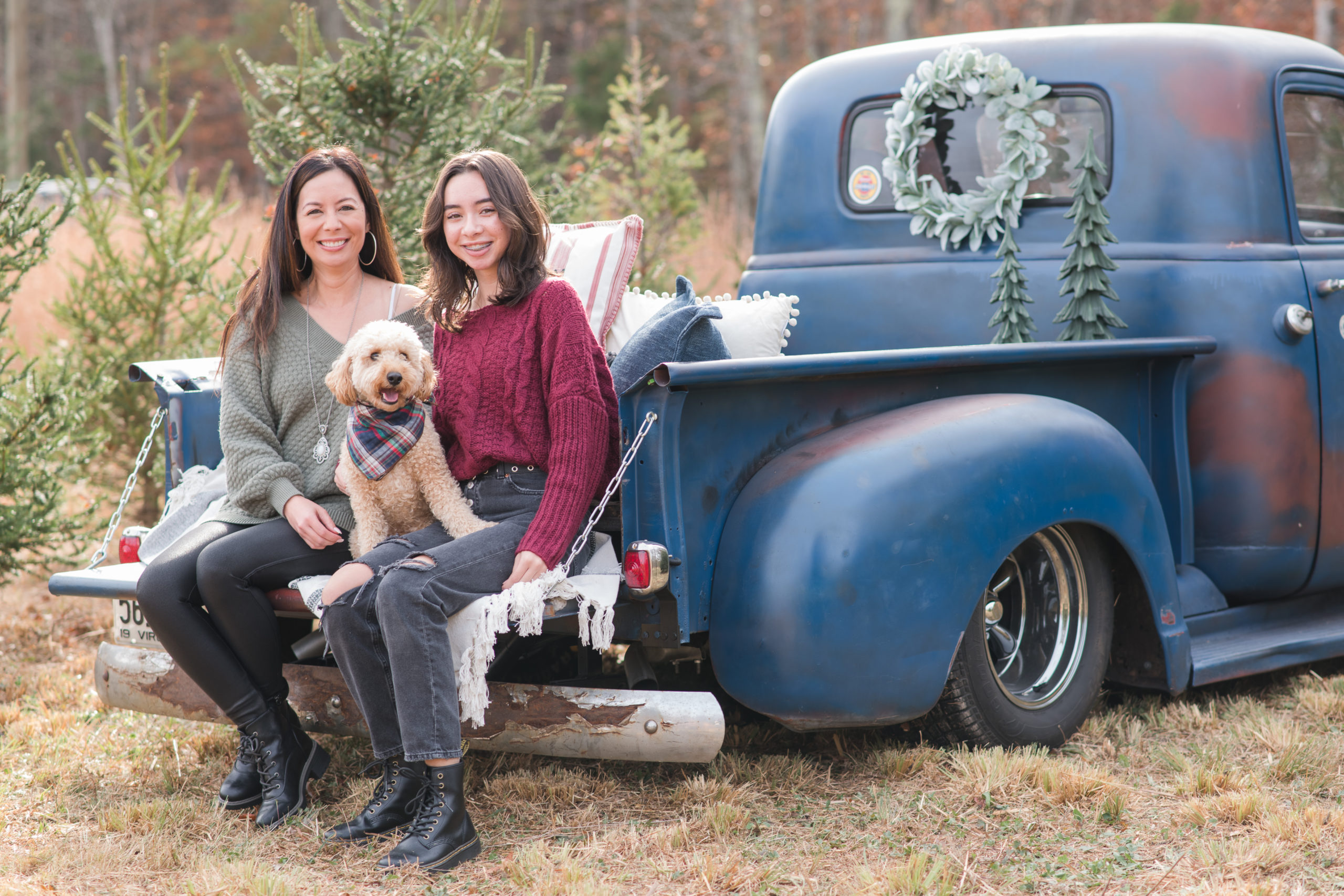 Mom and daughter with dog sitting on blue truck in christmas tree farm.