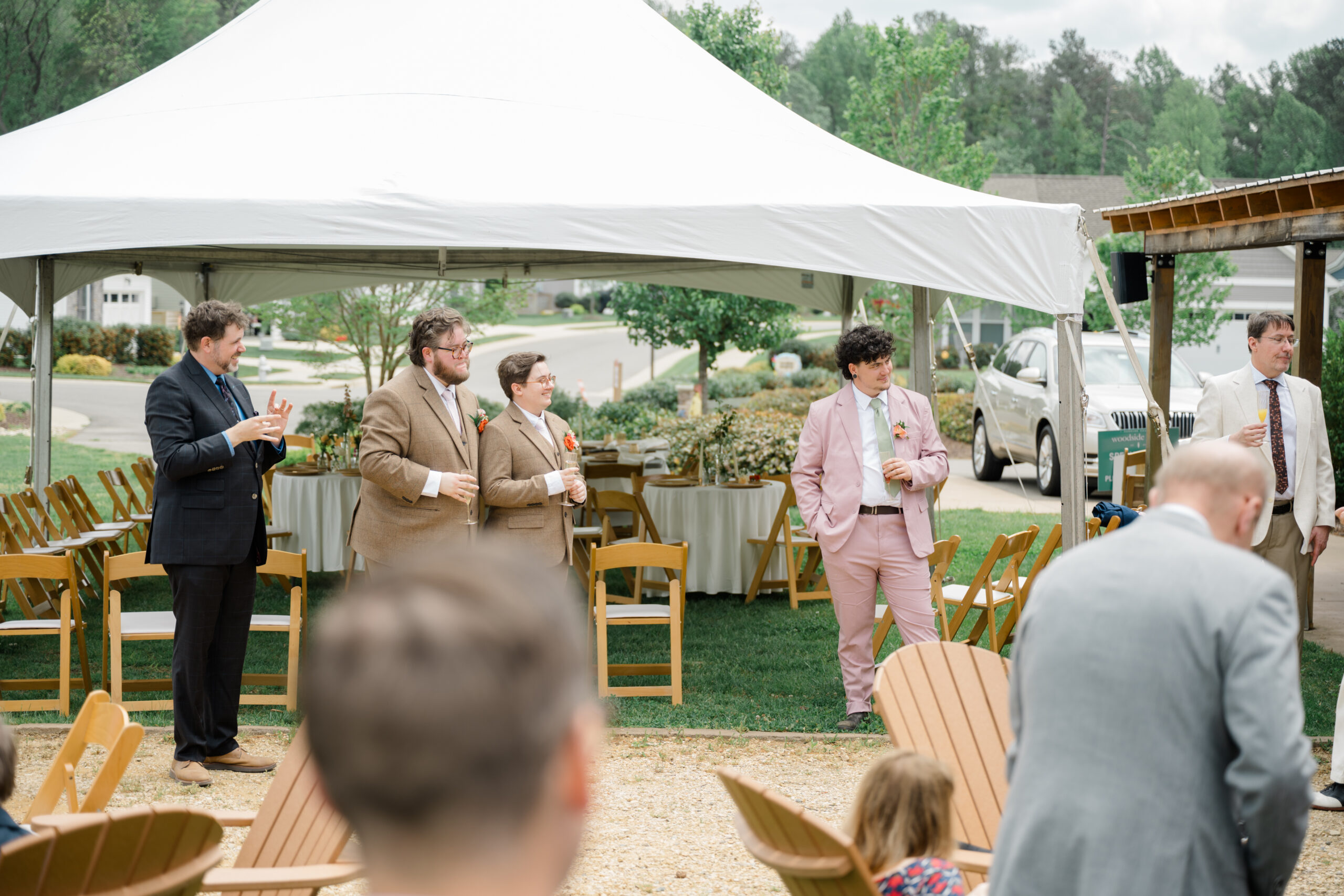 Two grooms stand with their wedding party during a toast while a guest uses American Sign Language to include all attendees in the celebration.
