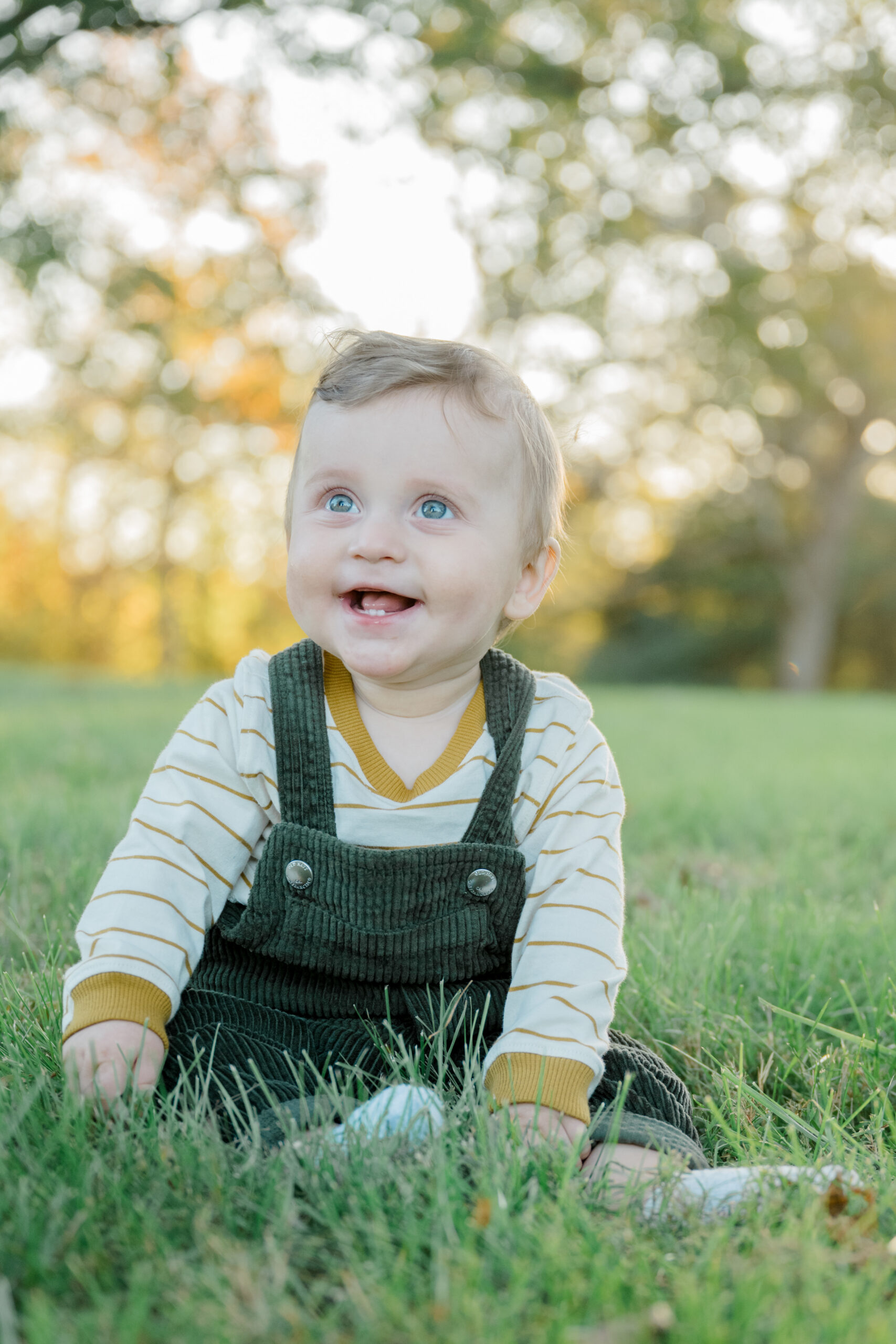 Baby boy with bright blue eyes sits in the grass wearing green corduroy overalls, captured during golden hour at a fall family session in Central Virginia.