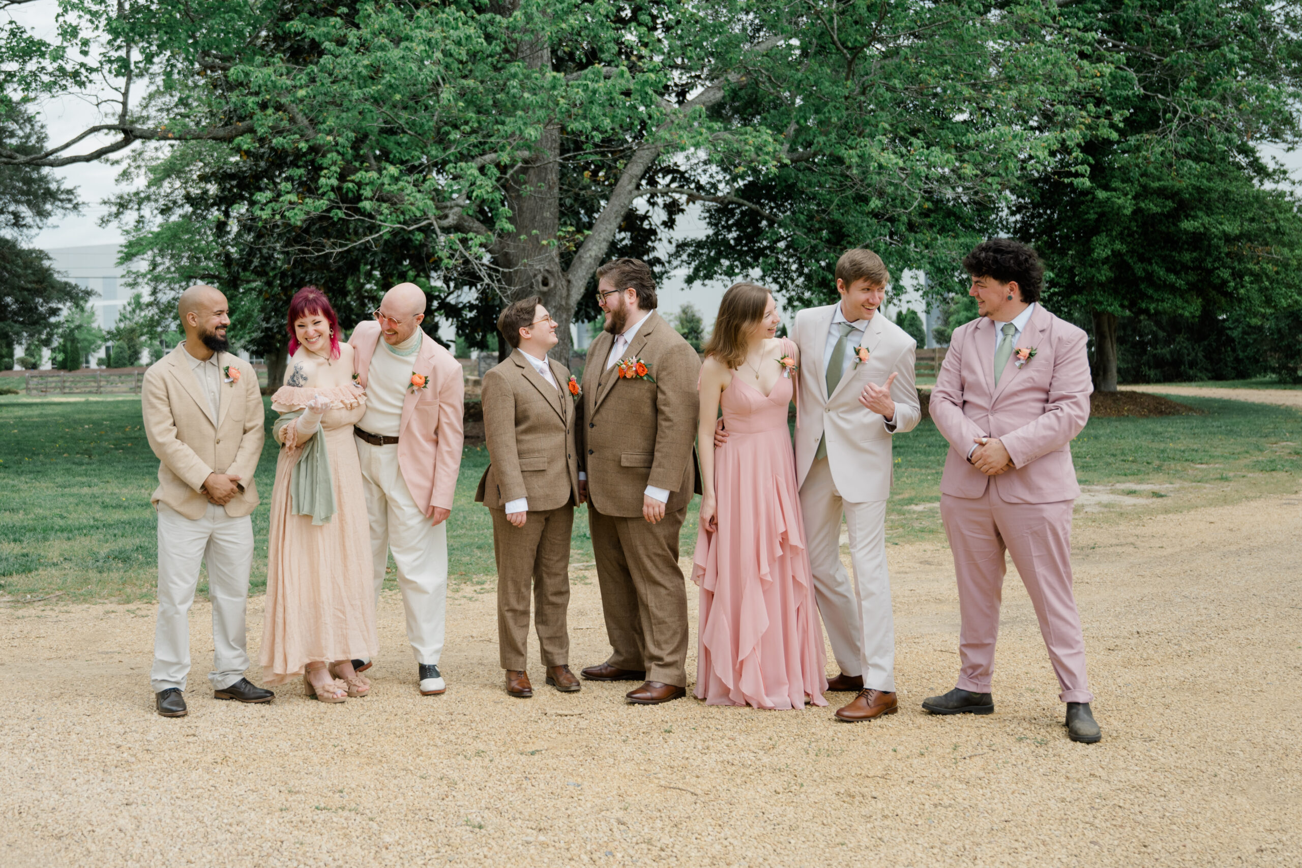 Full bridal party lineup for an LGBTQ+ wedding at Woodside Farms, with members in soft pink, cream, and tan suits and dresses standing together outdoors under tall trees.