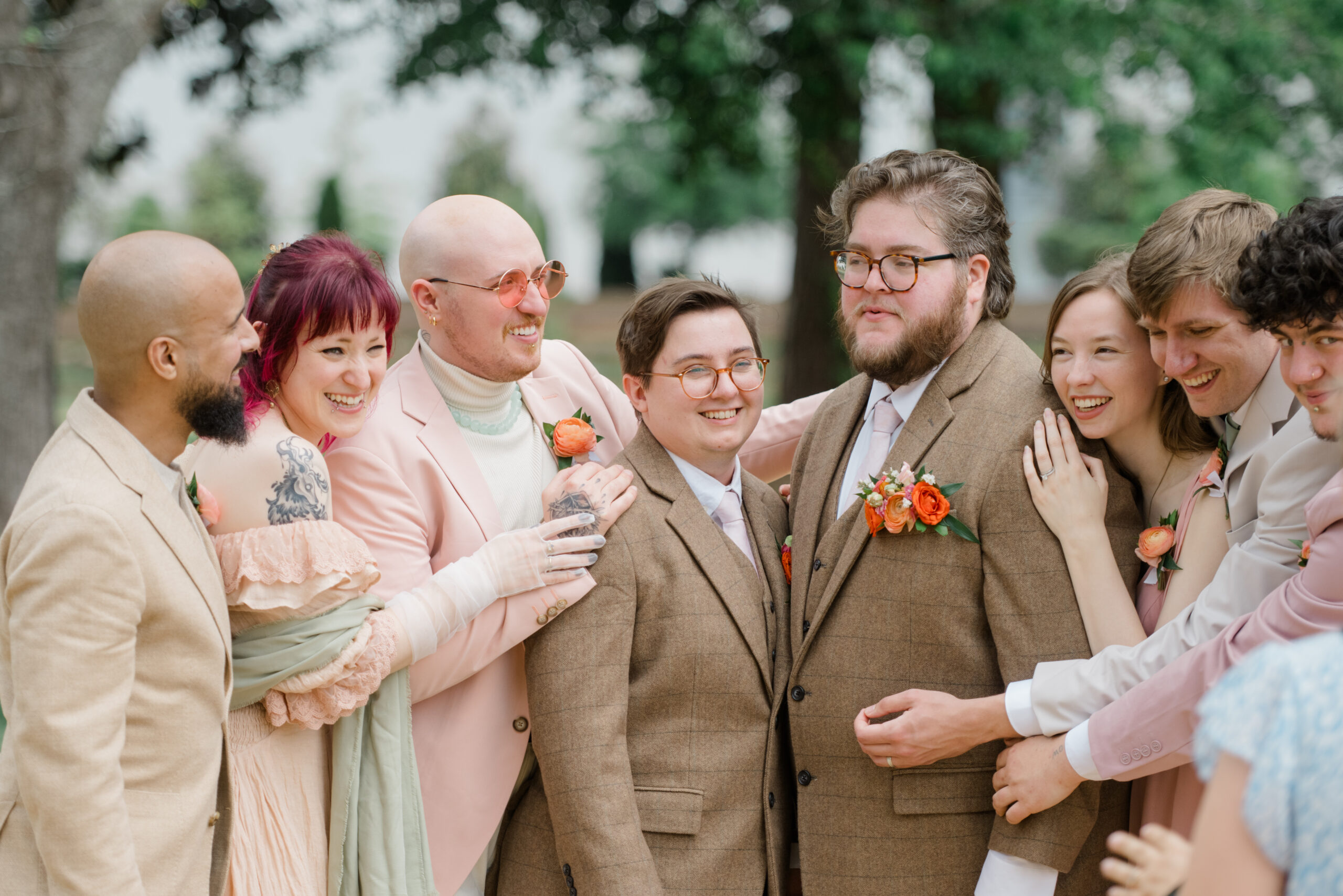 Joyful bridal party at a same-sex wedding laughing and embracing, with members wearing coordinated pastel and earth-toned outfits, including light pink, tan, and brown suits and dresses.