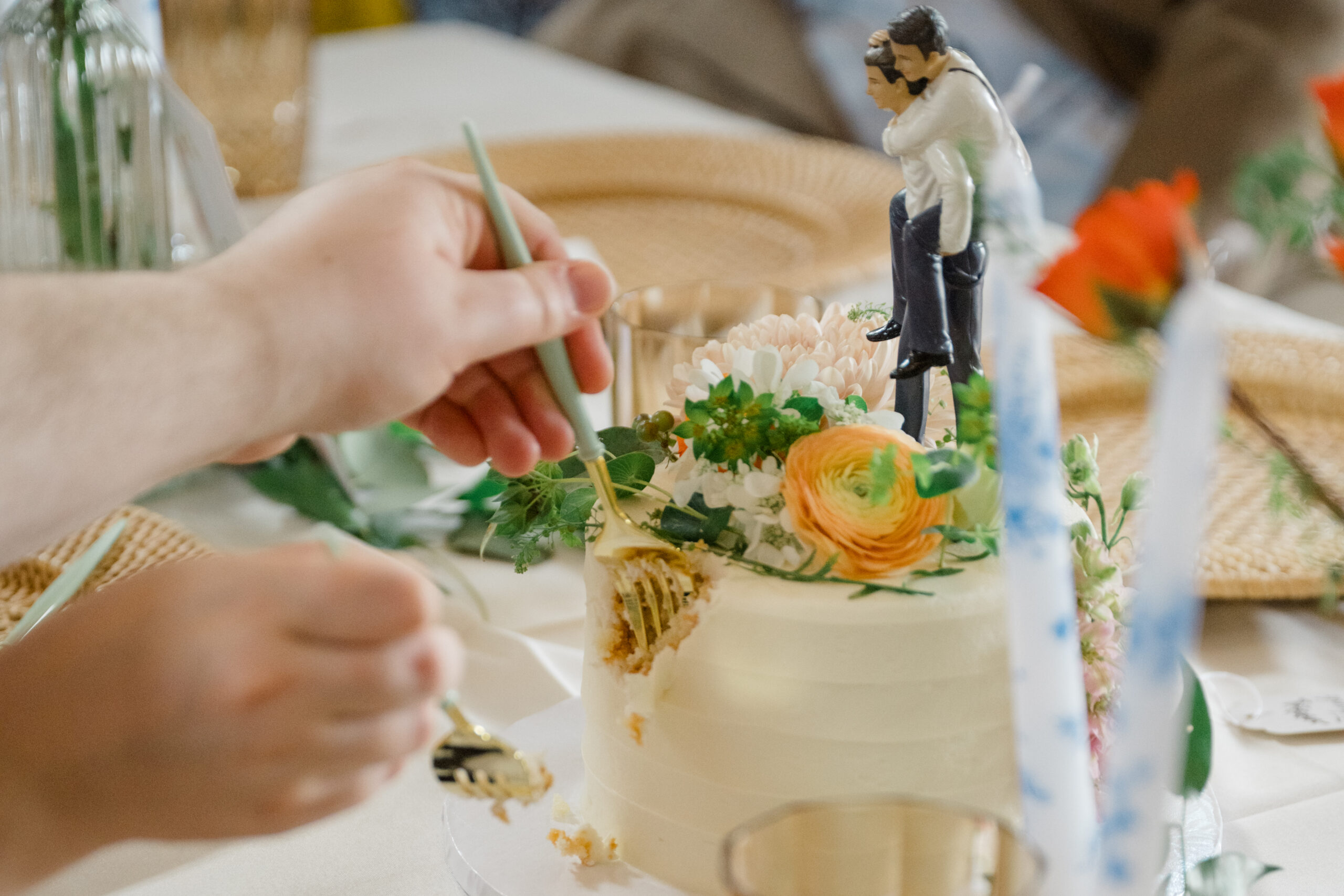 Detail shot of the same cake topper with grooms on a piggyback ride, surrounded by fresh flowers atop a white frosted cake.