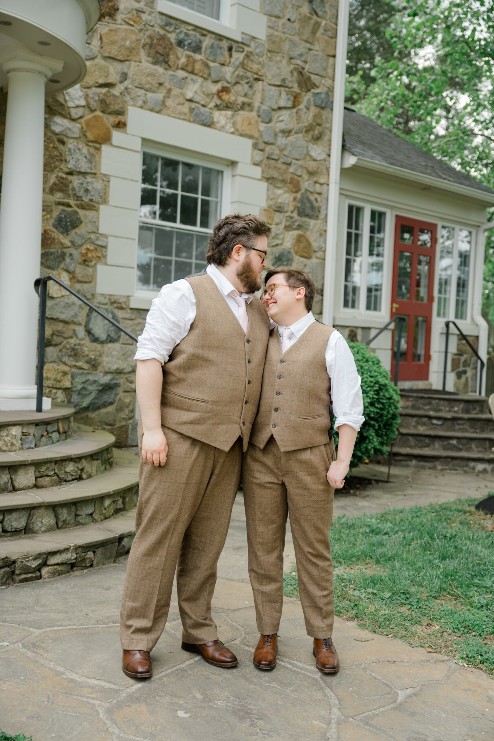 Two grooms in brown suits share an intimate moment, standing outside a stone manor house at Woodside Farms before their wedding ceremony.