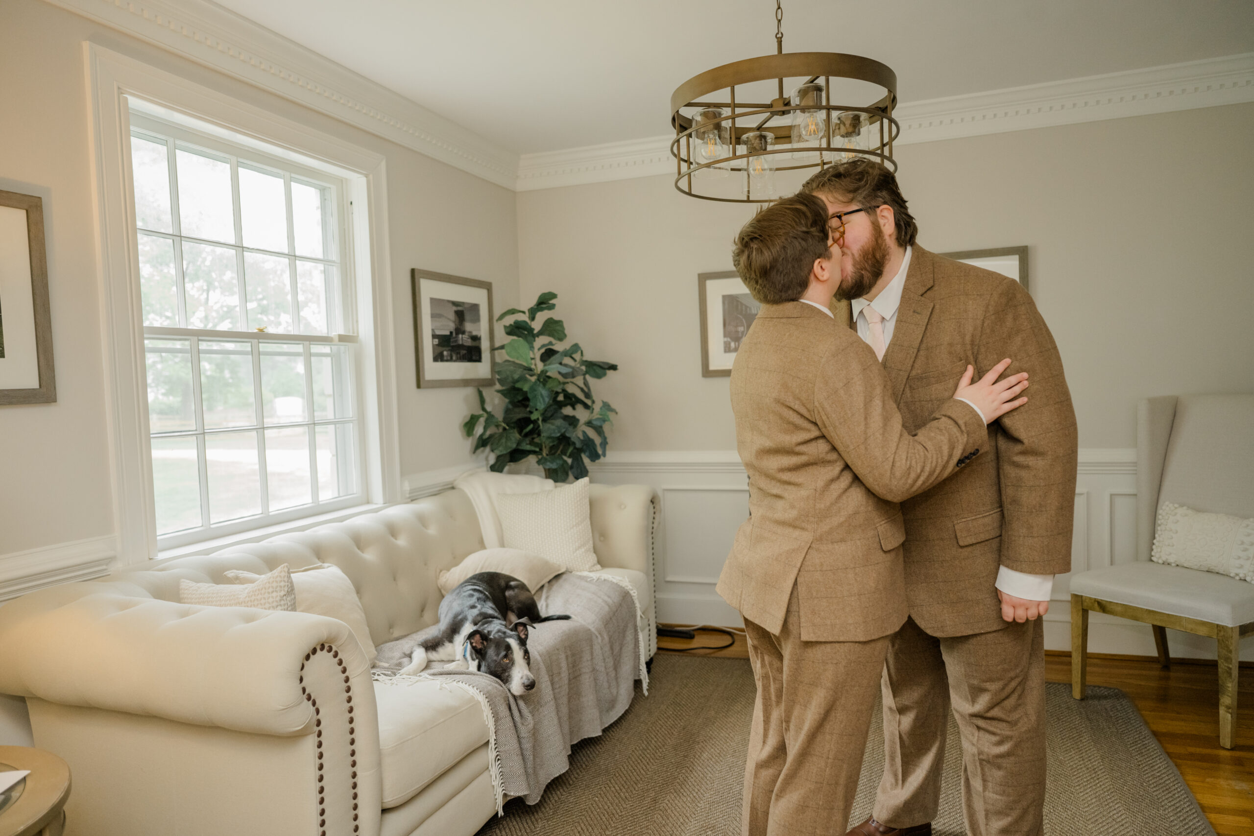 Two grooms share a kiss in their living room while their dog relaxes on the sofa behind them.