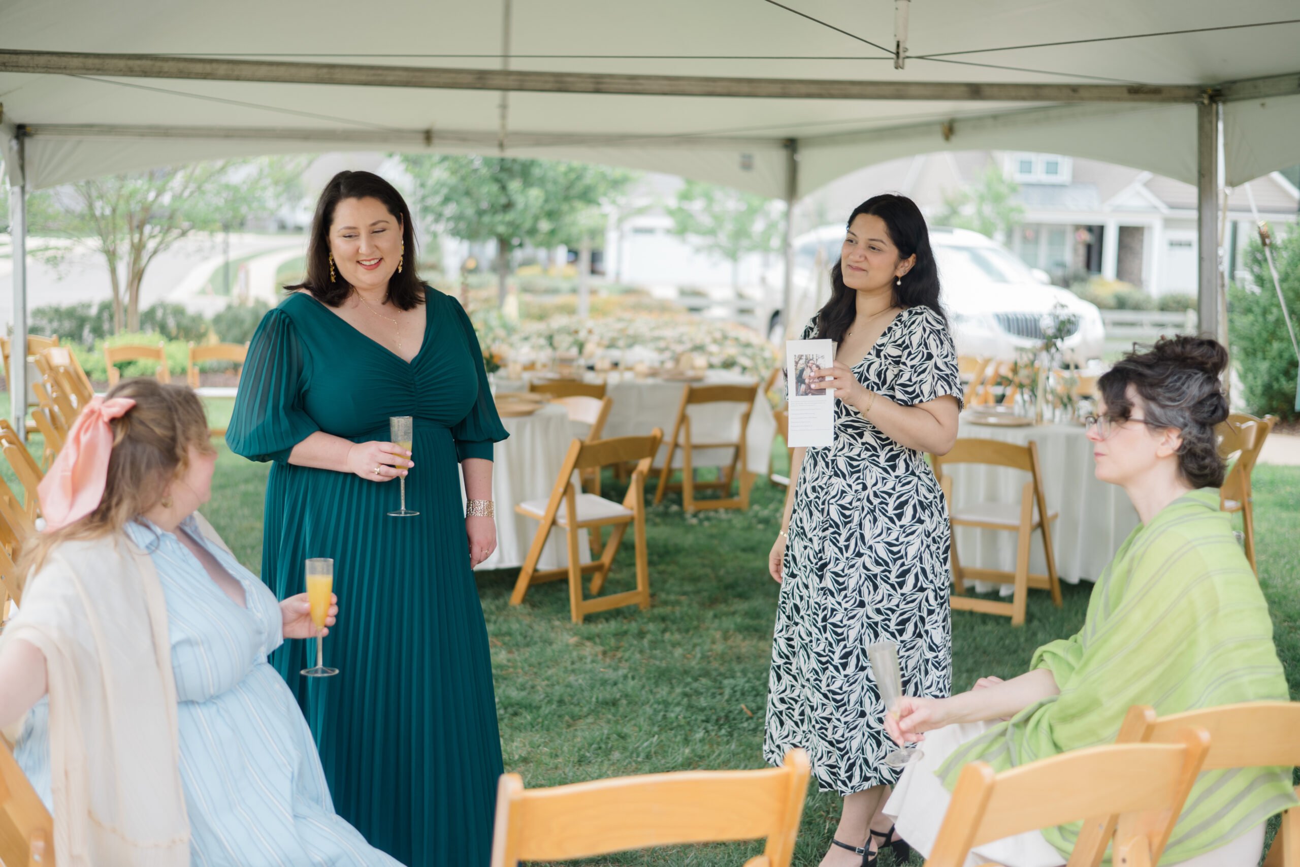 Guests chat and smile while holding mimosas under a white tent, surrounded by wooden chairs and set tables at a brunch wedding.