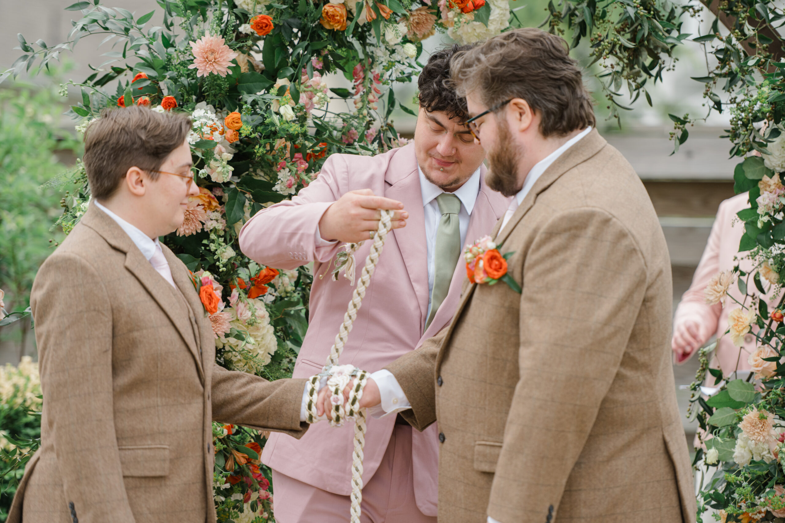 A bridesmaid in a blush pink dress helps tie handfasting ribbons around the grooms’ joined hands beneath a lush floral arch at Woodside Farms.