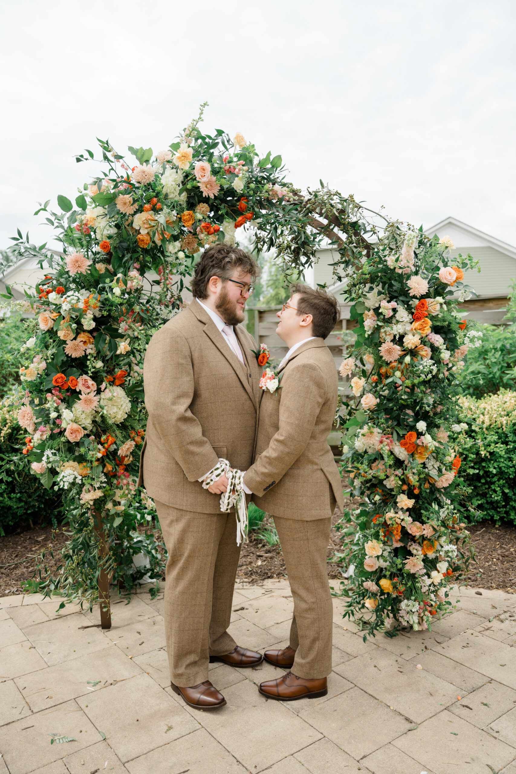 Two grooms stand hand-in-hand under a lush floral arch during their handfasting ceremony at Woodside Farms, wearing matching tan suits and smiling at one another with joy.