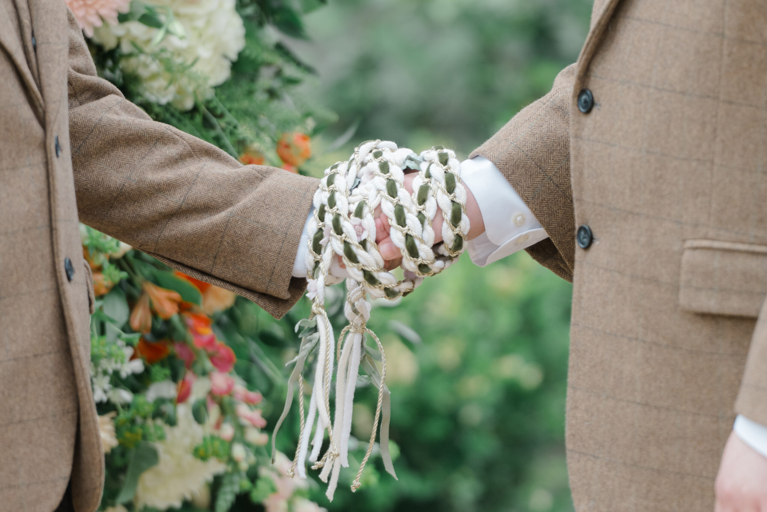 A close-up of intertwined hands wrapped in braided white and green handfasting ribbons during an LGBTQ+ wedding ceremony.