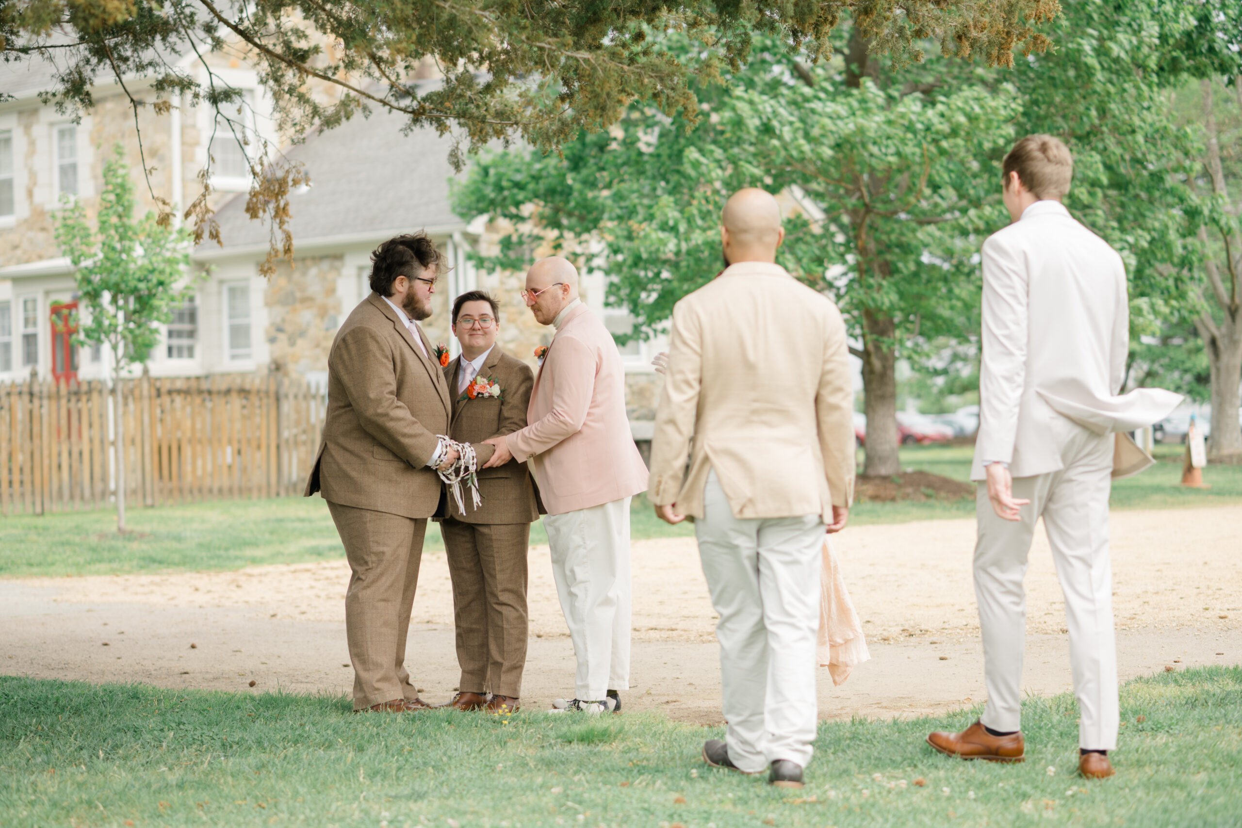 One of the grooms smiles while holding the intertwined handfasting ribbons after the ceremony, standing beside his husband under a canopy of trees.