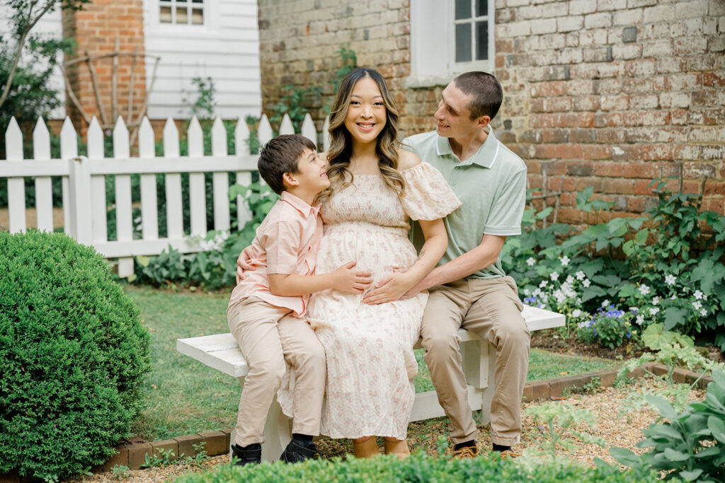 Smiling family sitting on a white bench during a summer maternity session at Historic Tuckahoe in Richmond, Virginia, featuring a pregnant mother in a floral dress with her partner and young son in a garden setting.