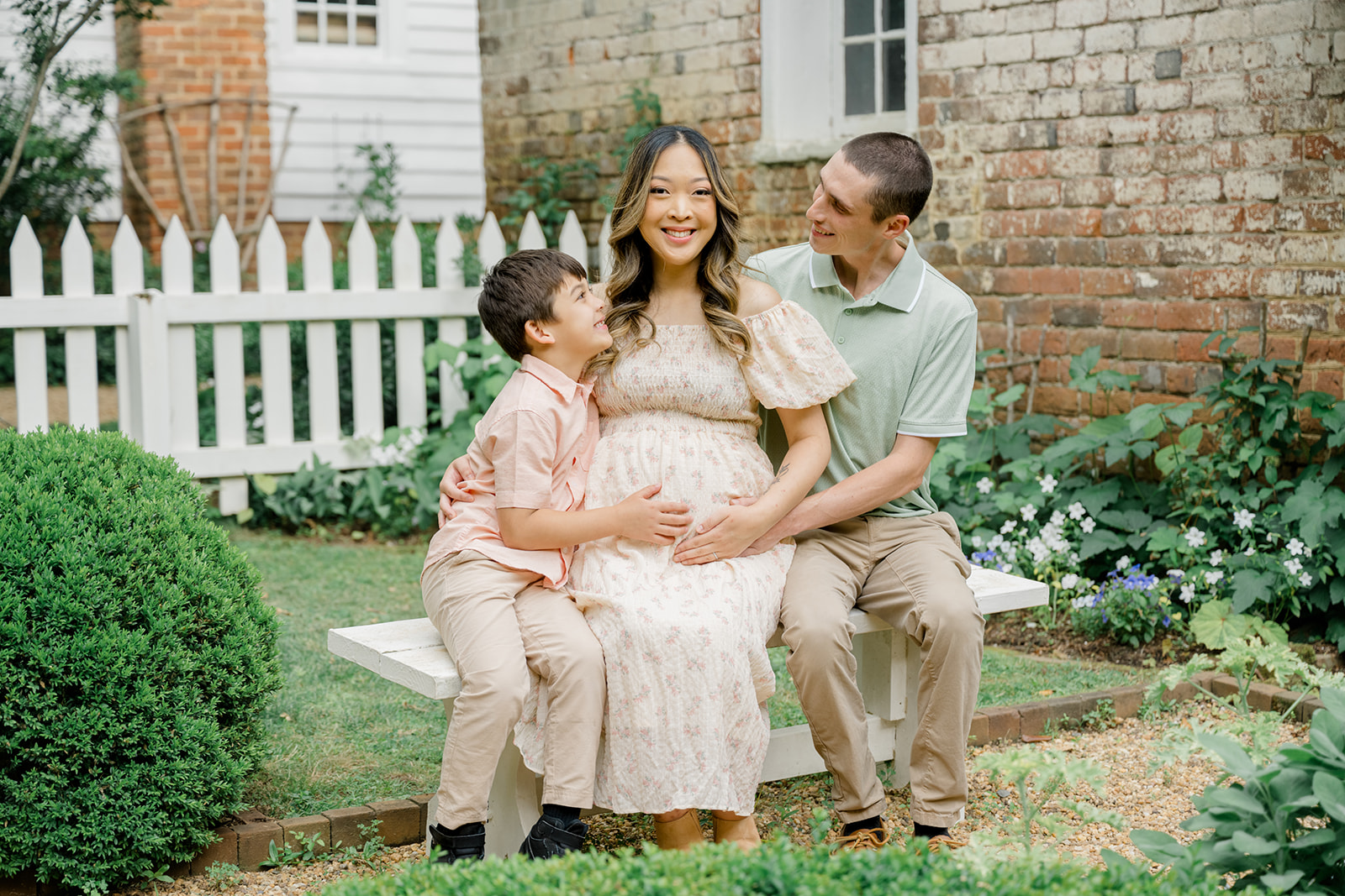 Smiling family sitting on a white bench during a summer maternity session at Historic Tuckahoe in Richmond, Virginia, featuring a pregnant mother in a floral dress with her partner and young son in a garden setting.