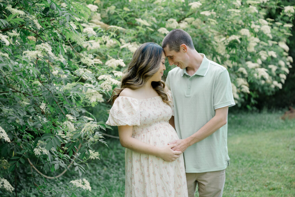 Intimate maternity photo of an expecting couple smiling with foreheads touching, surrounded by blooming white elderflowers at Historic Tuckahoe in Virginia.