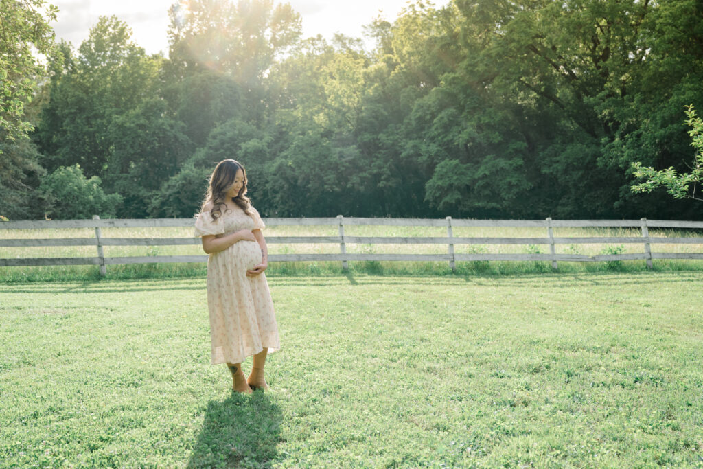 Solo maternity portrait of a mother in a floral dress standing barefoot in a sun-drenched field at golden hour at Historic Tuckahoe.
