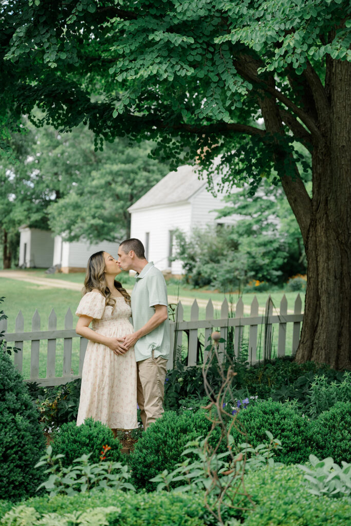 Pregnant couple kisses beneath a lush tree in the manicured gardens of Historic Tuckahoe during a romantic Virginia maternity photo session.
