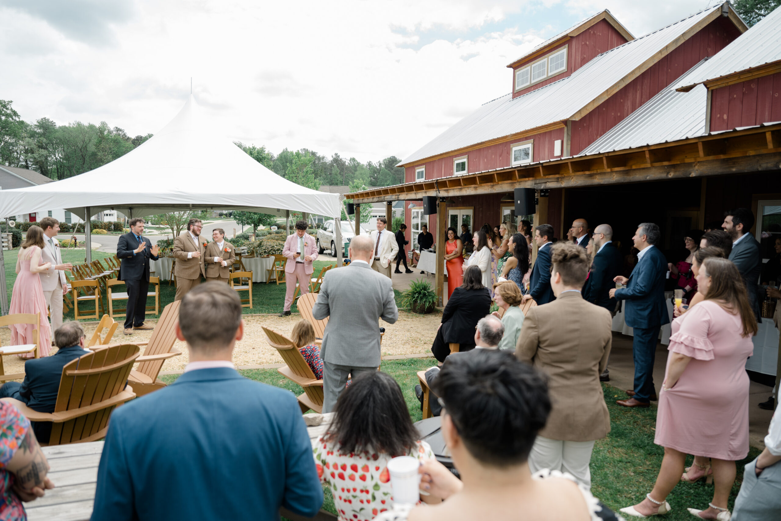 Wide view of an outdoor brunch wedding reception with guests gathered under a tent and near a red barn, listening to a toast in progress.