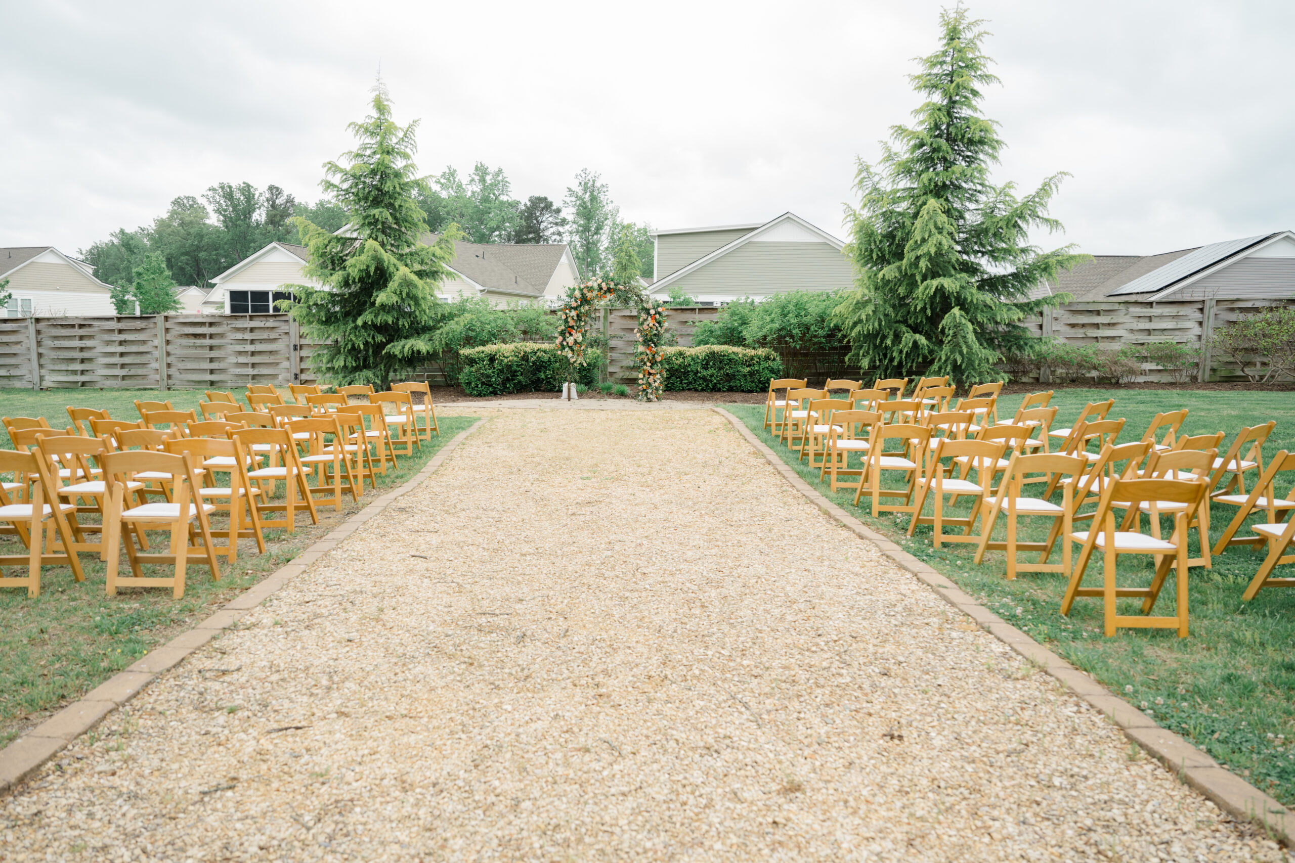 Outdoor wedding ceremony setup with rows of wooden chairs and a floral arch in front of a garden fence.
