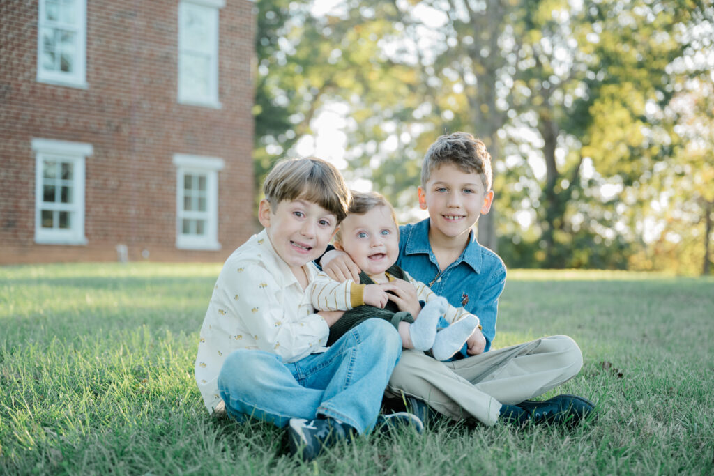 Three brothers sit closely together on the grass with their baby brother in the center, surrounded by soft golden light during their Virginia family portrait session.