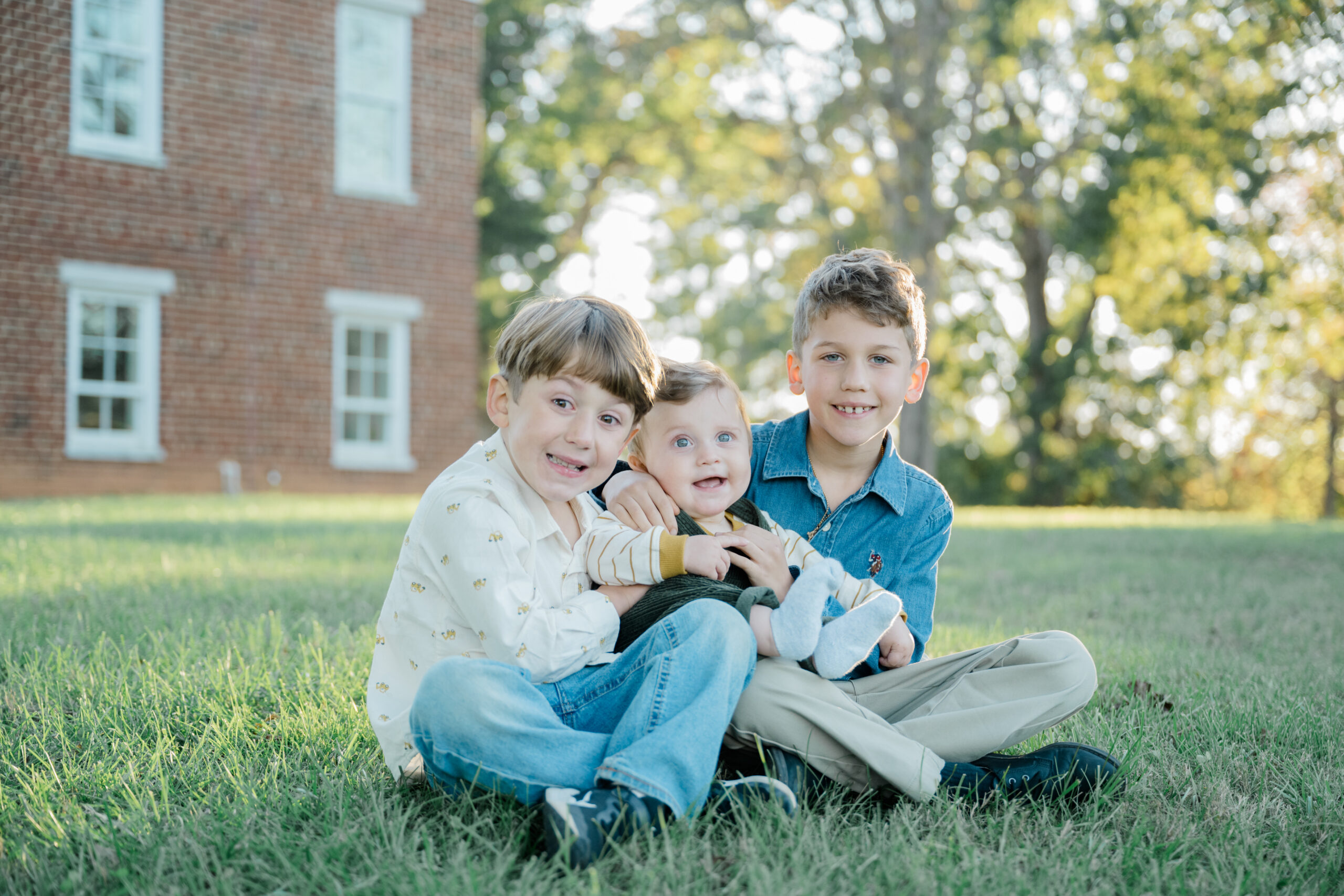 Three brothers sit closely together on the grass with their baby brother in the center, surrounded by soft golden light during their Virginia family portrait session.