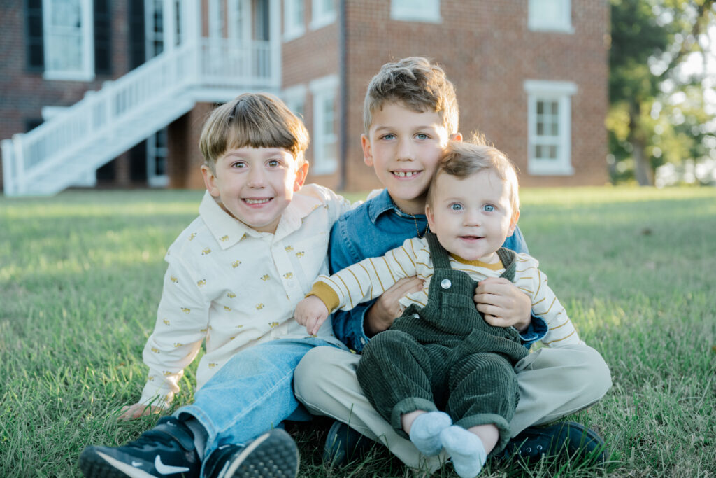 Three young brothers sit together on grass in front of a historic brick building during a fall family session at Pleasant Grove Park in Fluvanna, Virginia.
