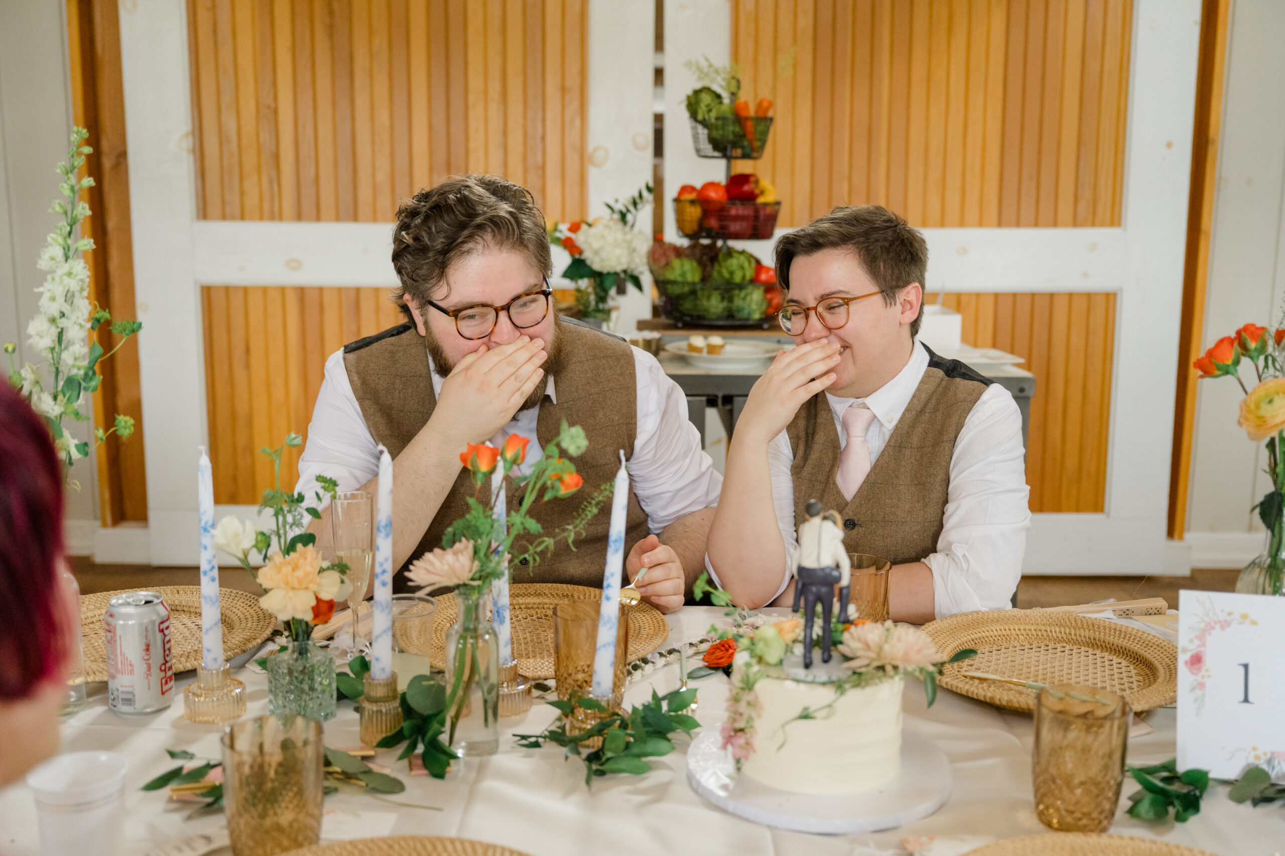 The grooms laugh together at their sweetheart table during the reception, surrounded by floral centerpieces and a wedding cake featuring their custom topper.