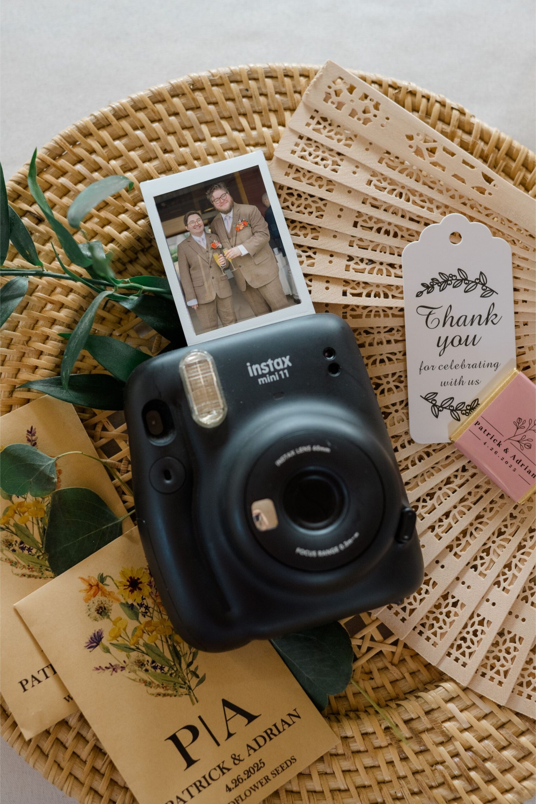 Flat lay of wedding details including an Instax camera with a printed photo of the grooms, seed packet favors, a thank-you tag, and a wooden fan—styled on a wicker charger plate.