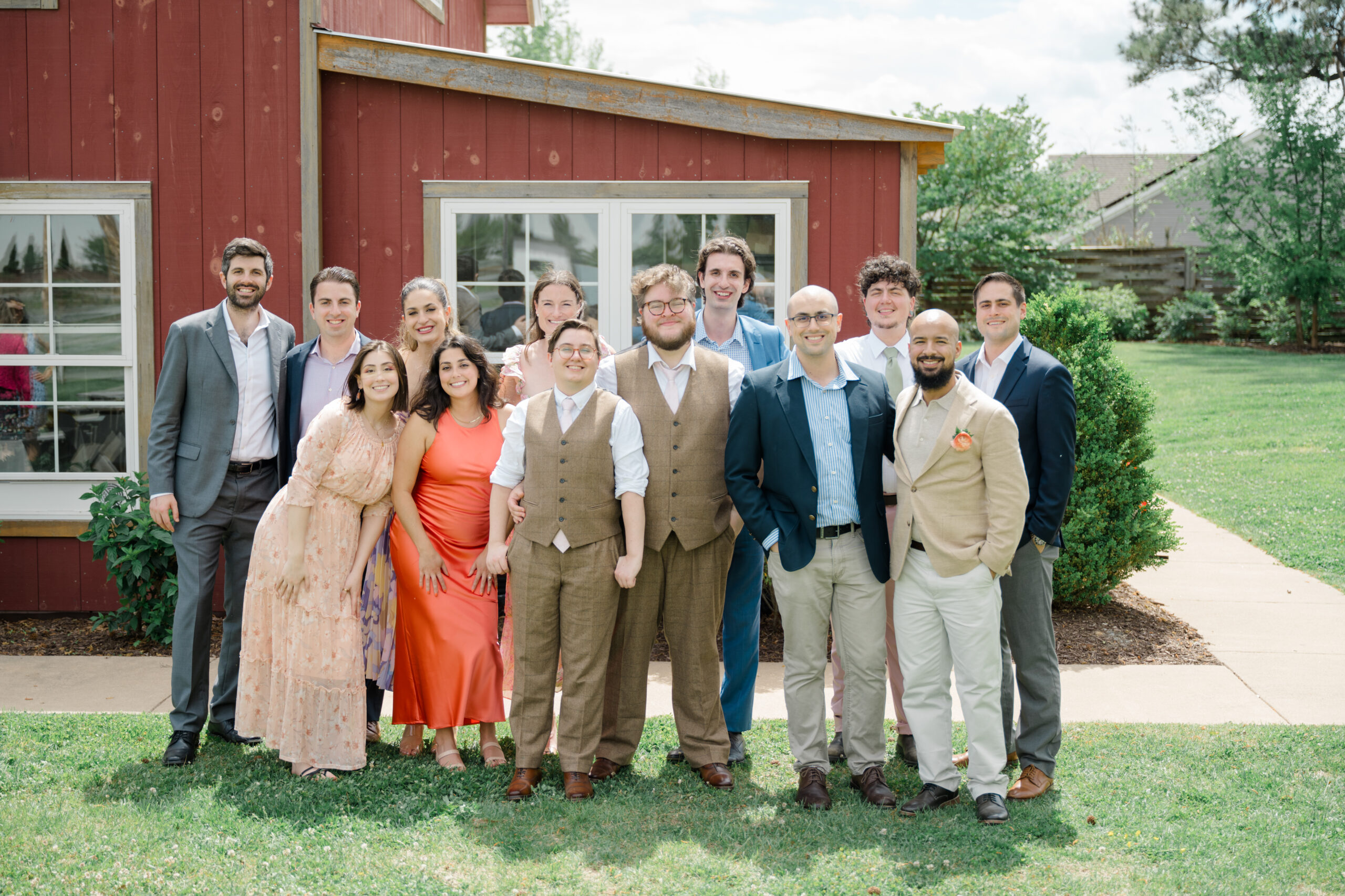 The grooms pose for a fun group photo with friends and loved ones, all smiling and making playful gestures in front of a red barn.