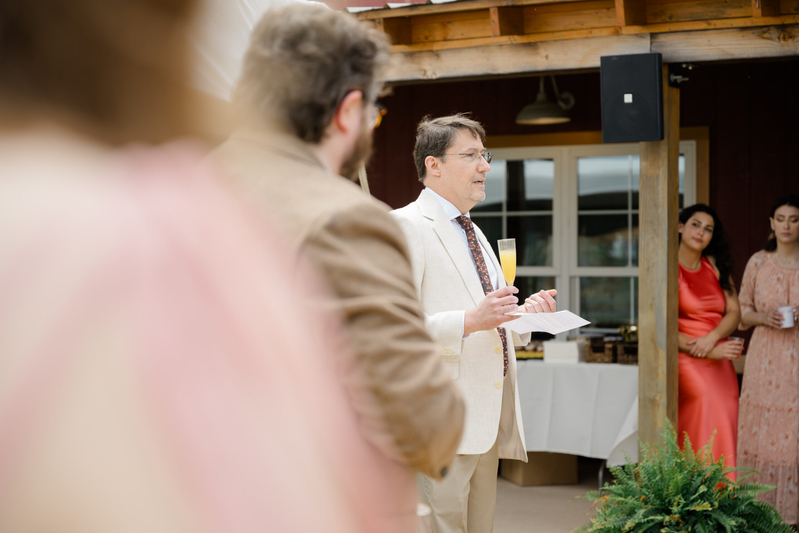 Man in a cream suit holding a mimosa and giving a wedding toast outside a barn at a brunch reception, with wedding guests listening nearby.