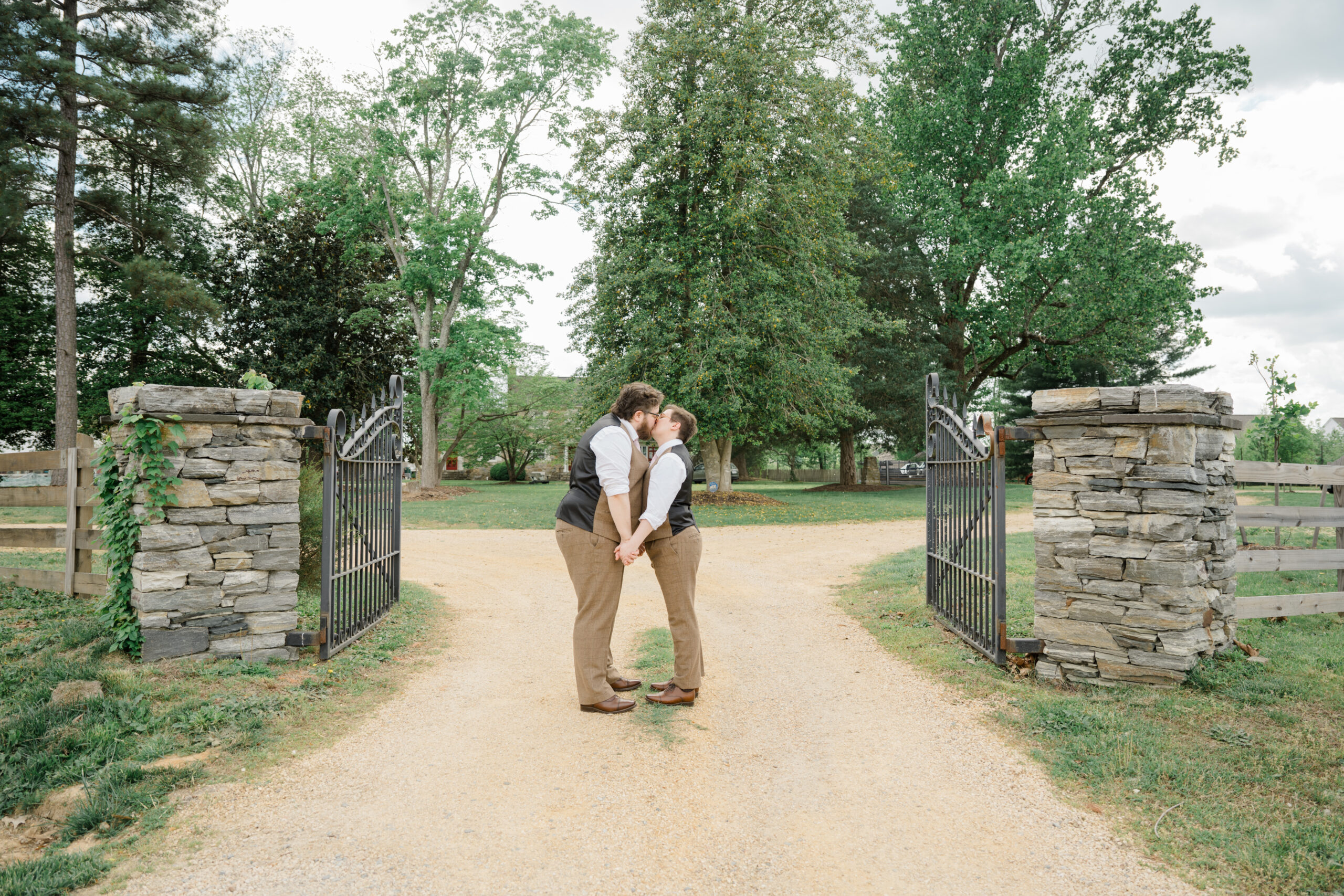 Two grooms sharing a kiss in front of an open iron gate at Woodside Farms, standing on a gravel path surrounded by greenery and mature trees.
