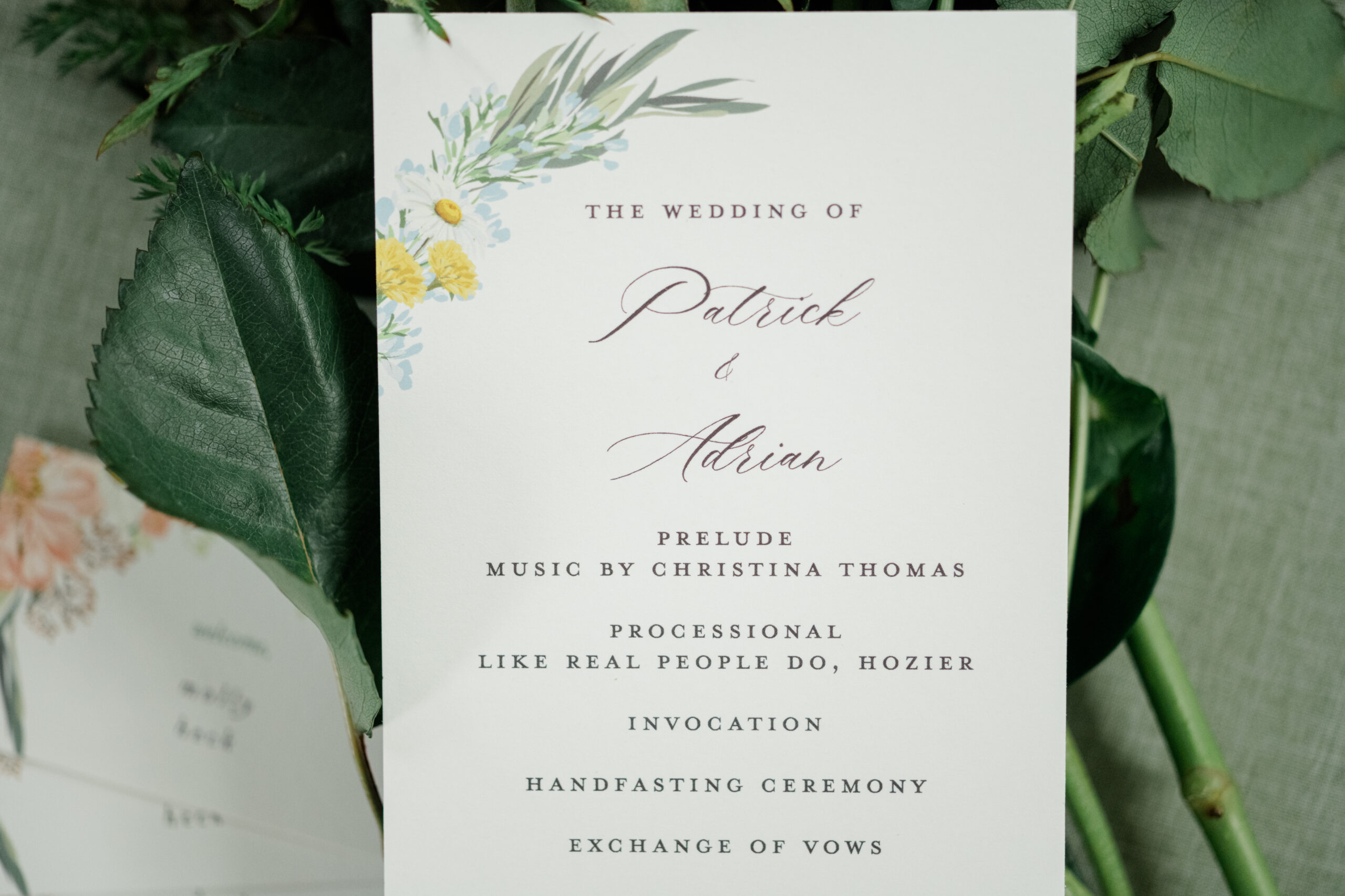 Close-up of the spring wedding ceremony program for Patrick and Adrian, nestled among greenery and florals.