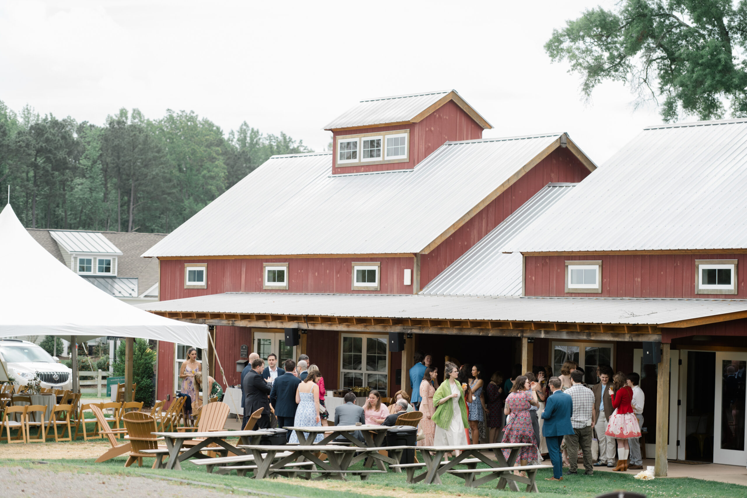 Wide shot of the red barn venue at Woodside Farms during a brunch wedding reception, with guests mingling under a white tent and on picnic benches outside.