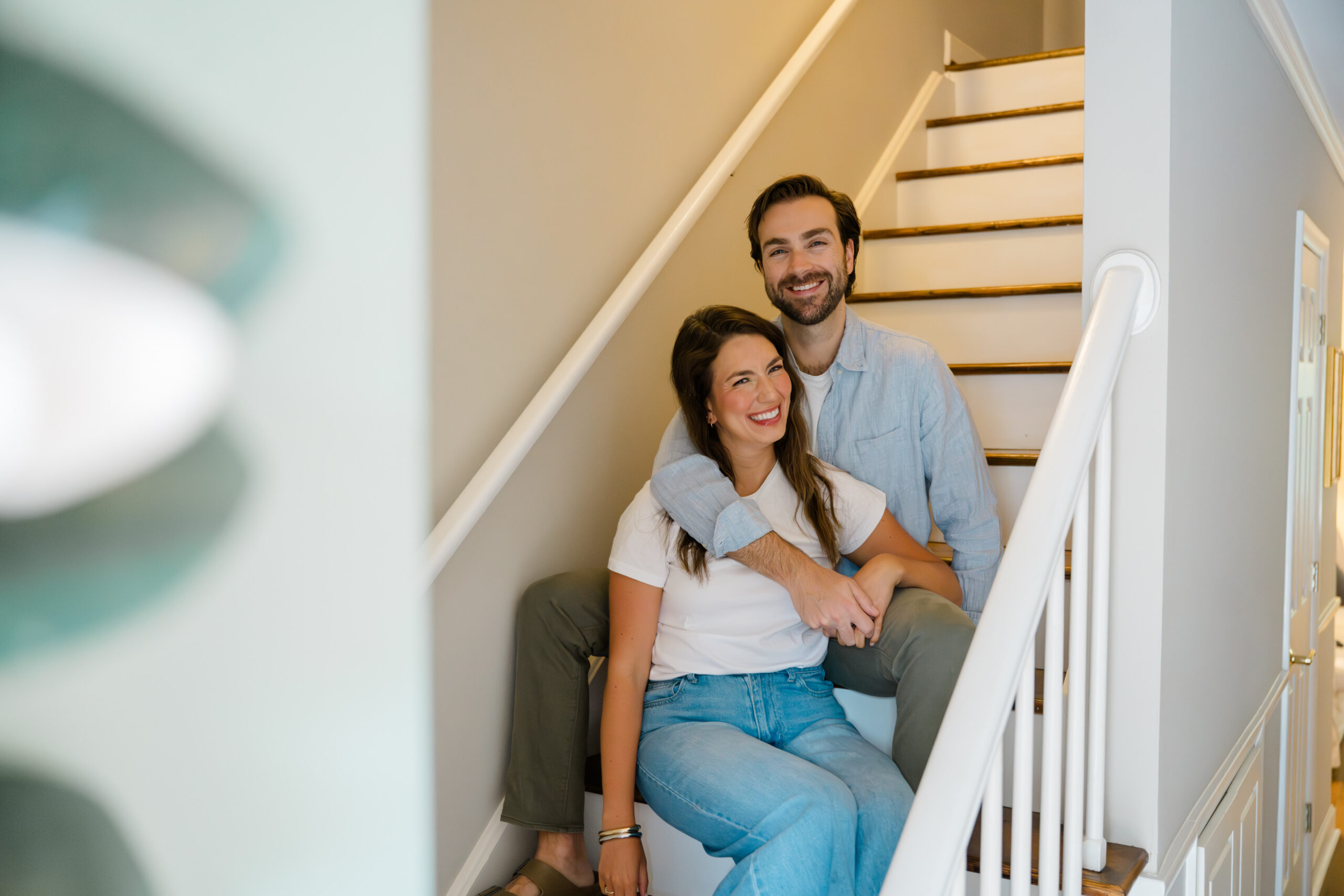 Couple sitting together on a staircase, laughing and wrapped in each other’s arms—sunny, relaxed, and completely at ease in their home.
