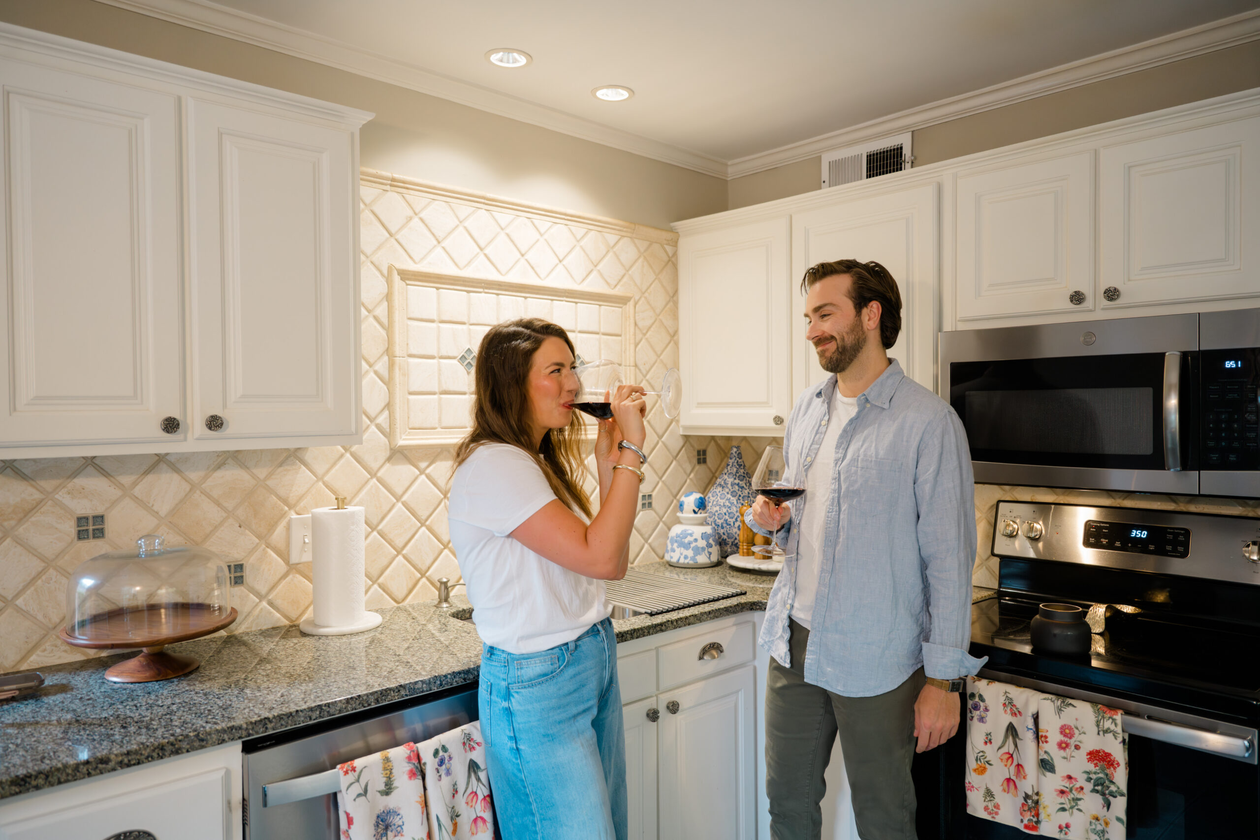 Couple enjoying red wine together in a cozy, warmly lit kitchen, smiling and relaxed as they lean against the counter surrounded by charming floral accents and neutral tile backsplash.
