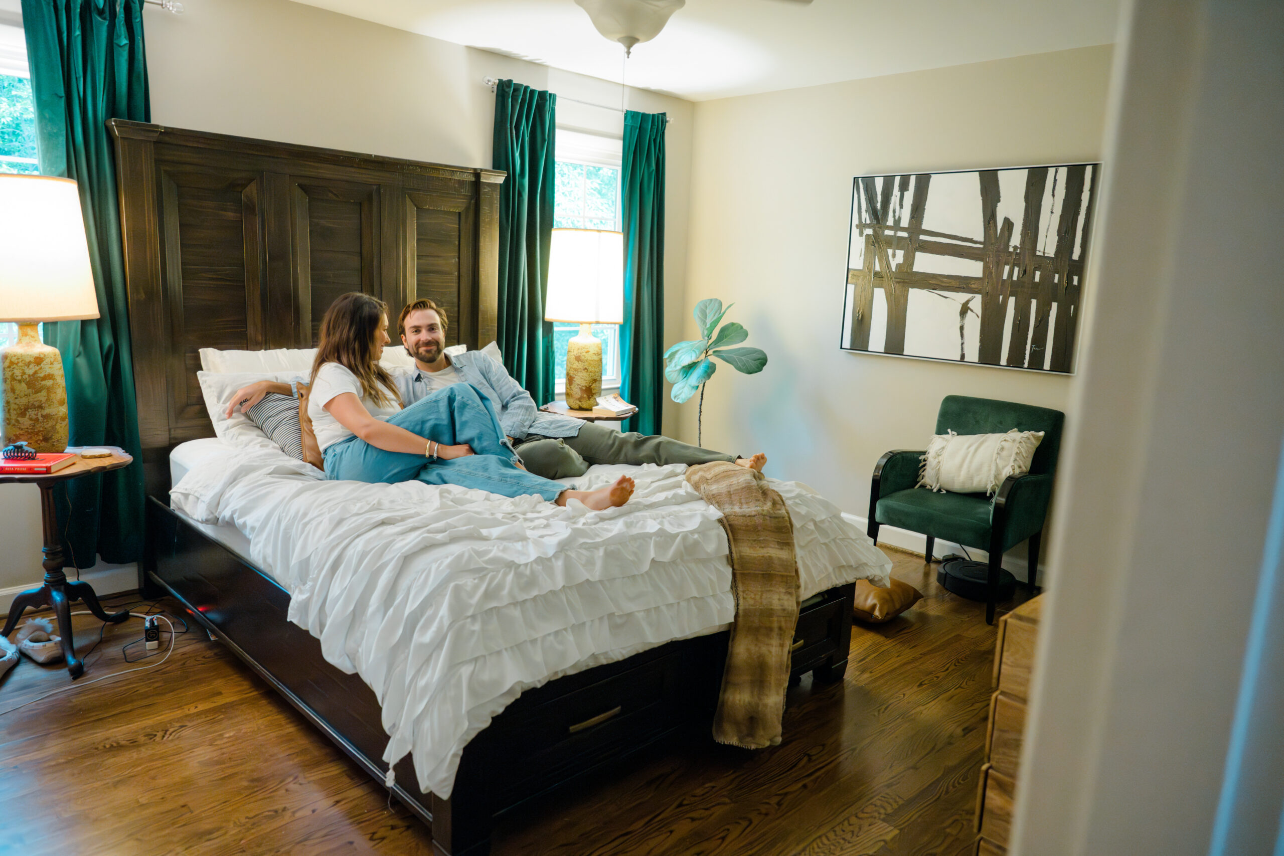 Couple lounging together on a neatly made bed in a warmly decorated bedroom, surrounded by emerald green curtains, golden light from matching lamps, and cozy modern decor.
