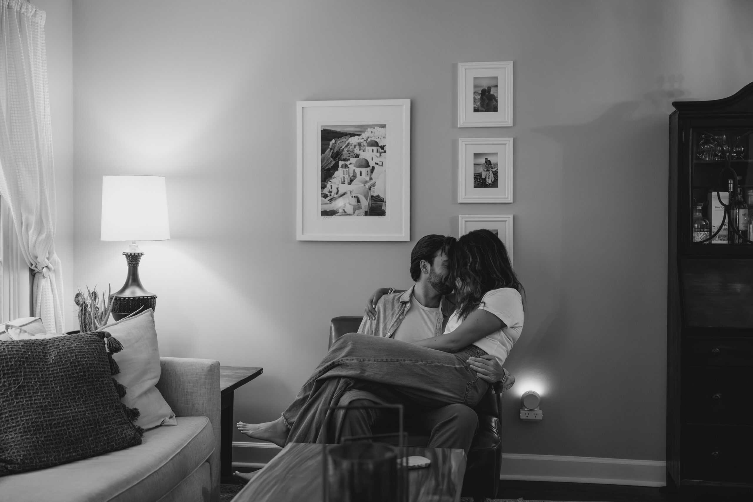 Black-and-white photo of a couple embracing on a cozy armchair in their Charlotte living room, surrounded by framed memories and soft evening light during their in-home documentary photo session.
