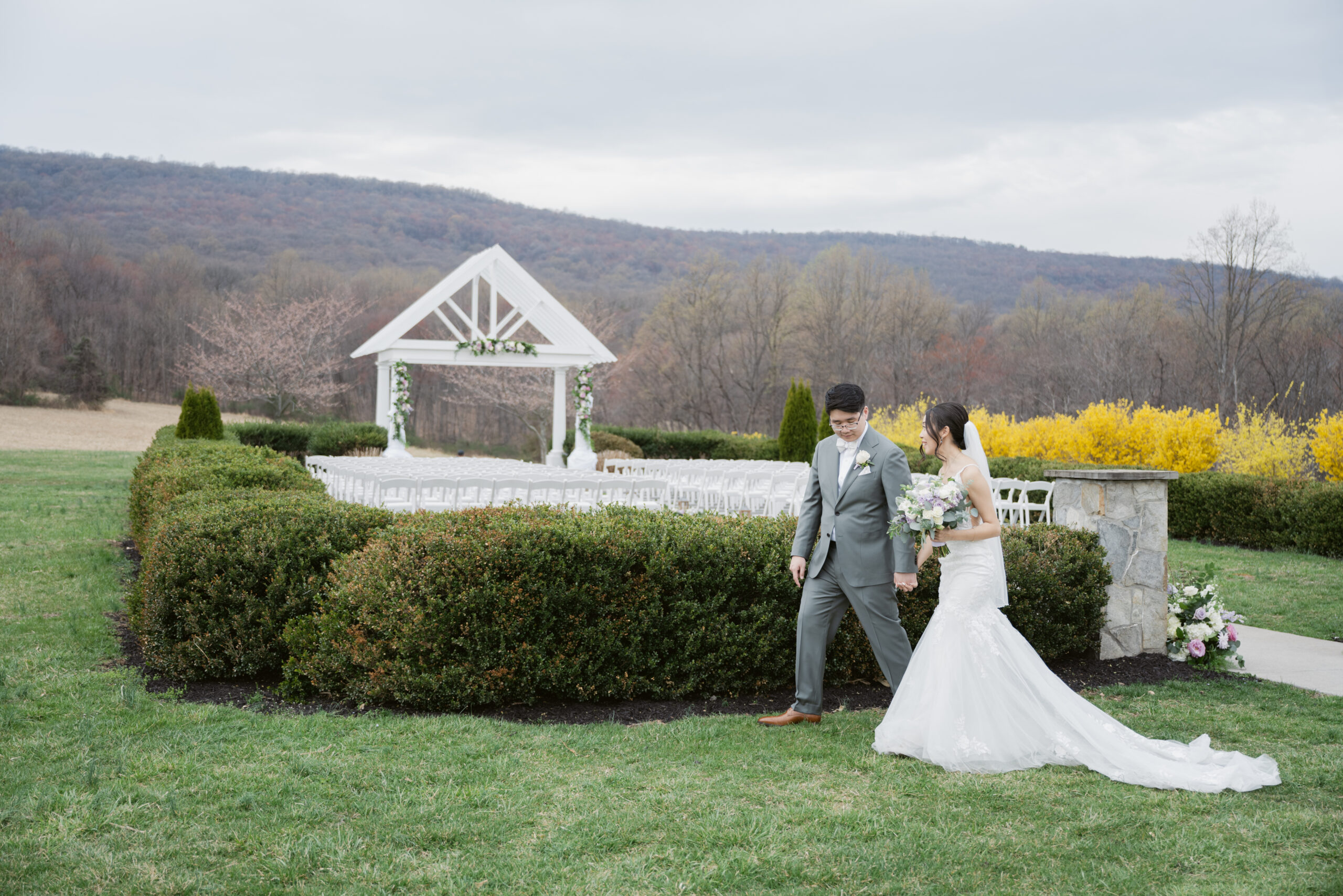 Bride and groom walking hand in hand near a white ceremony arbor with mountain views at Springfield Manor, surrounded by spring greenery and yellow blooms.