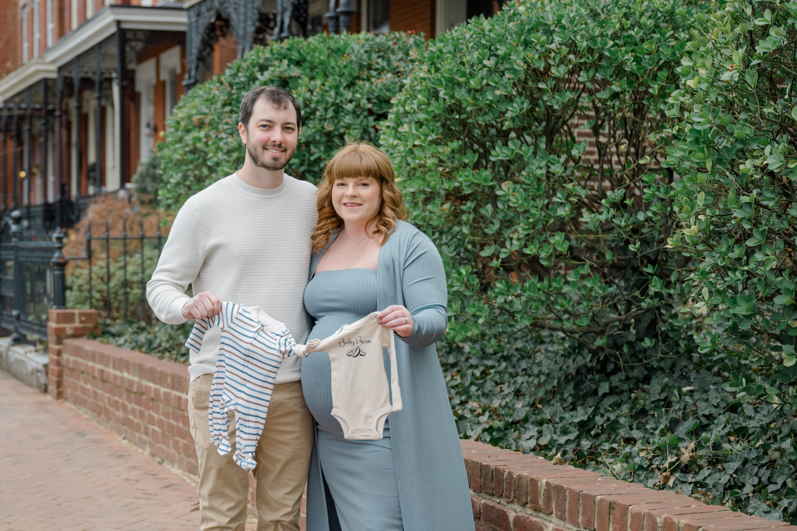 Expecting couple smiling while holding up baby onesies, standing in front of a historic row of homes in Richmond during their late February maternity session.
