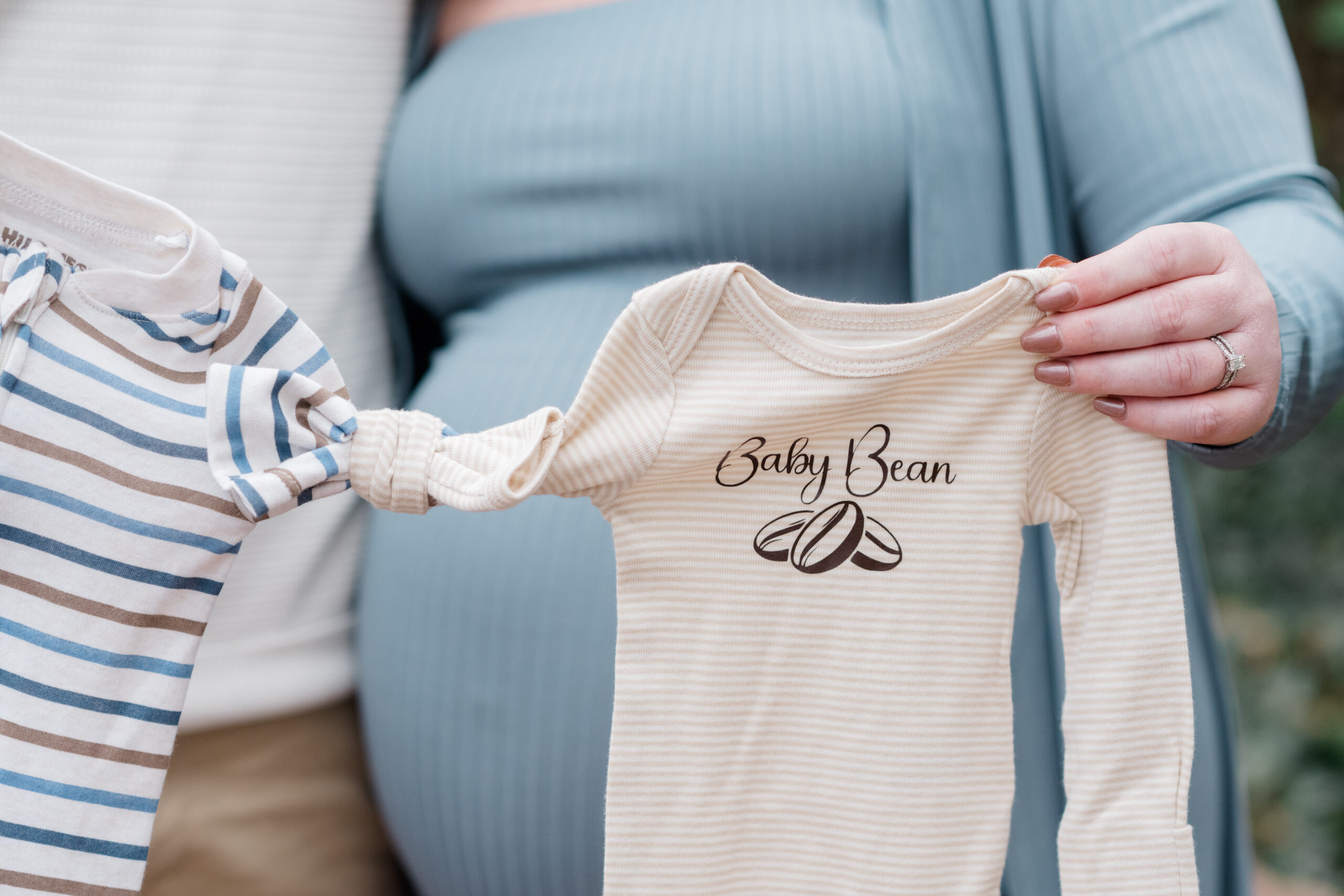 Close-up of a beige baby onesie that says “Baby Bean,” held by expecting parents in coordinating blue and white outfits during a maternity session at Libby Hill Park in Richmond, VA.