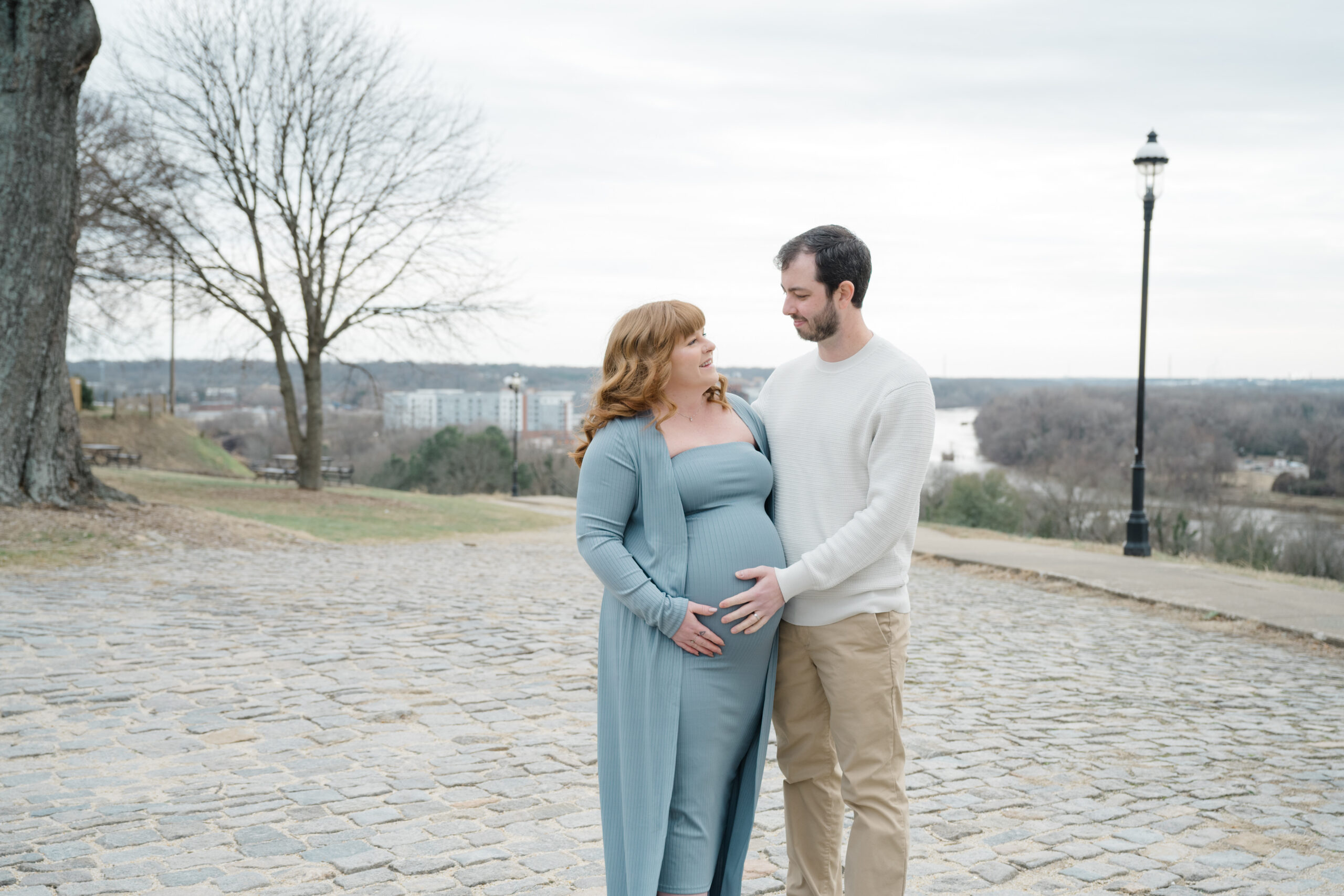 Parents-to-be embracing on cobblestone streets at Libby Hill Park with scenic views of downtown Richmond in the background.