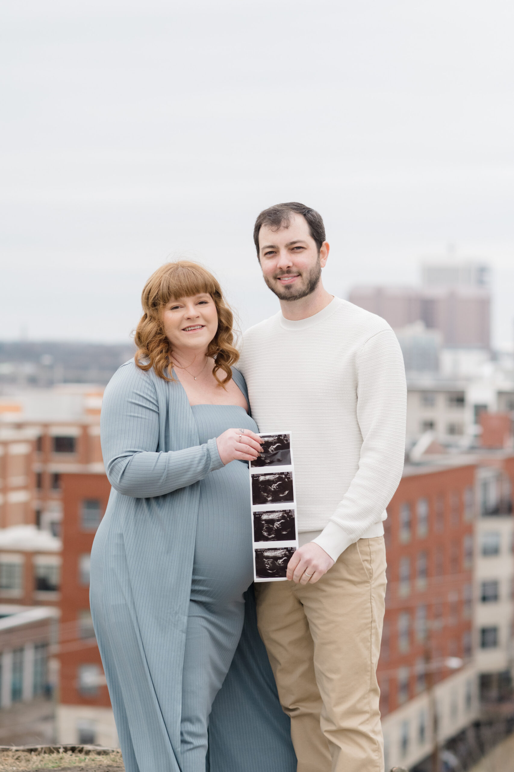 Couple standing together on a hill holding their baby’s ultrasound photos with the Richmond city skyline behind them.