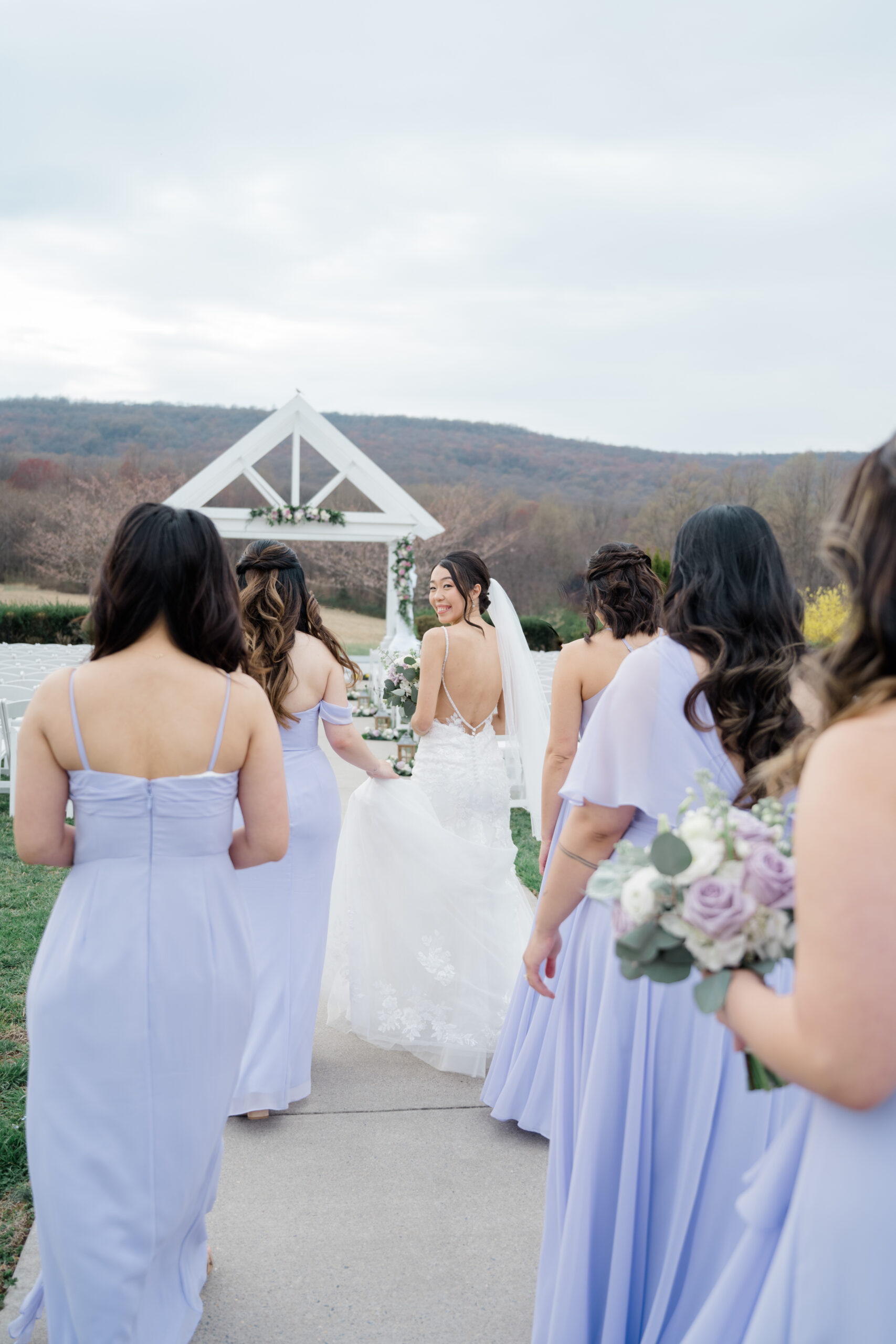 Bride walking with her bridesmaids in lavender dresses toward a white ceremony arbor.