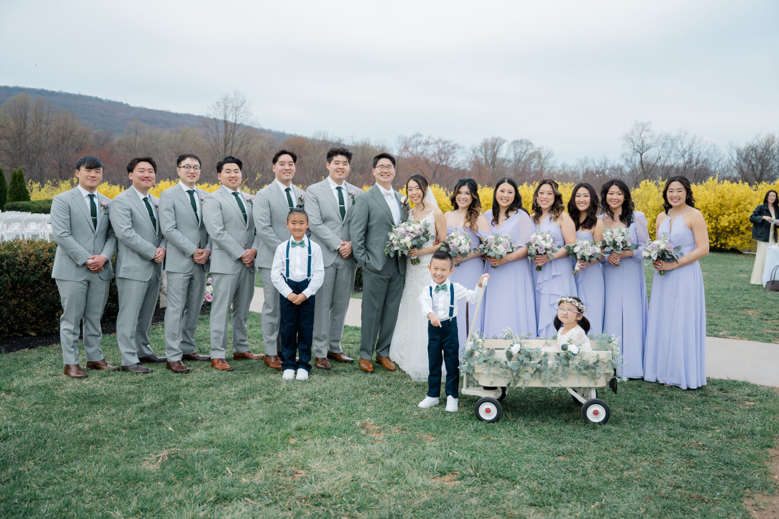 Bride and groom posing with their full wedding party at Springfield Manor, including groomsmen in light gray suits, bridesmaids in lavender dresses, two ring bearers in suspenders, and a flower girl seated in a white floral-decorated wagon with spring blooms in the background.