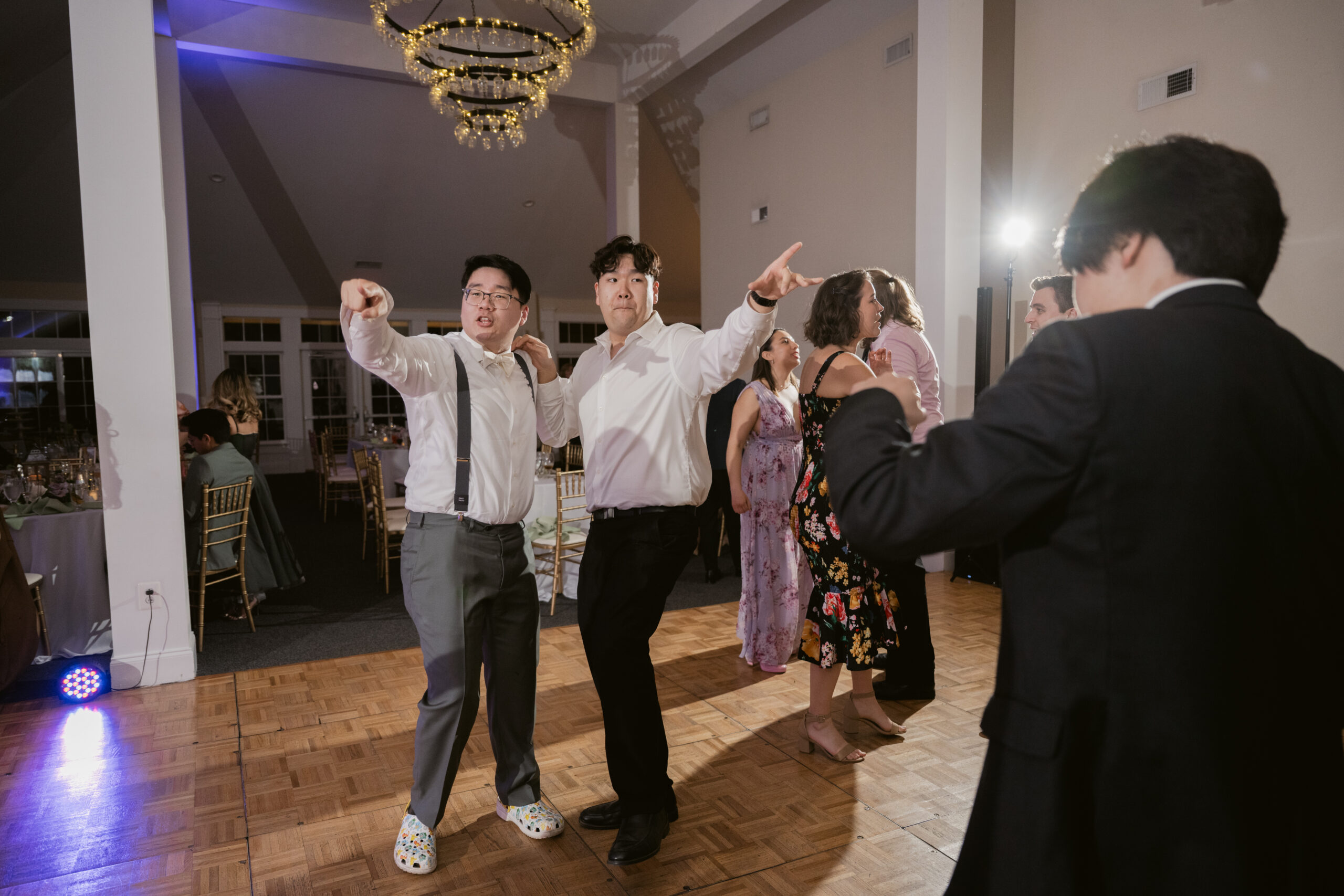 Two groomsmen dancing playfully and pointing while guests dance in the background under a chandelier.