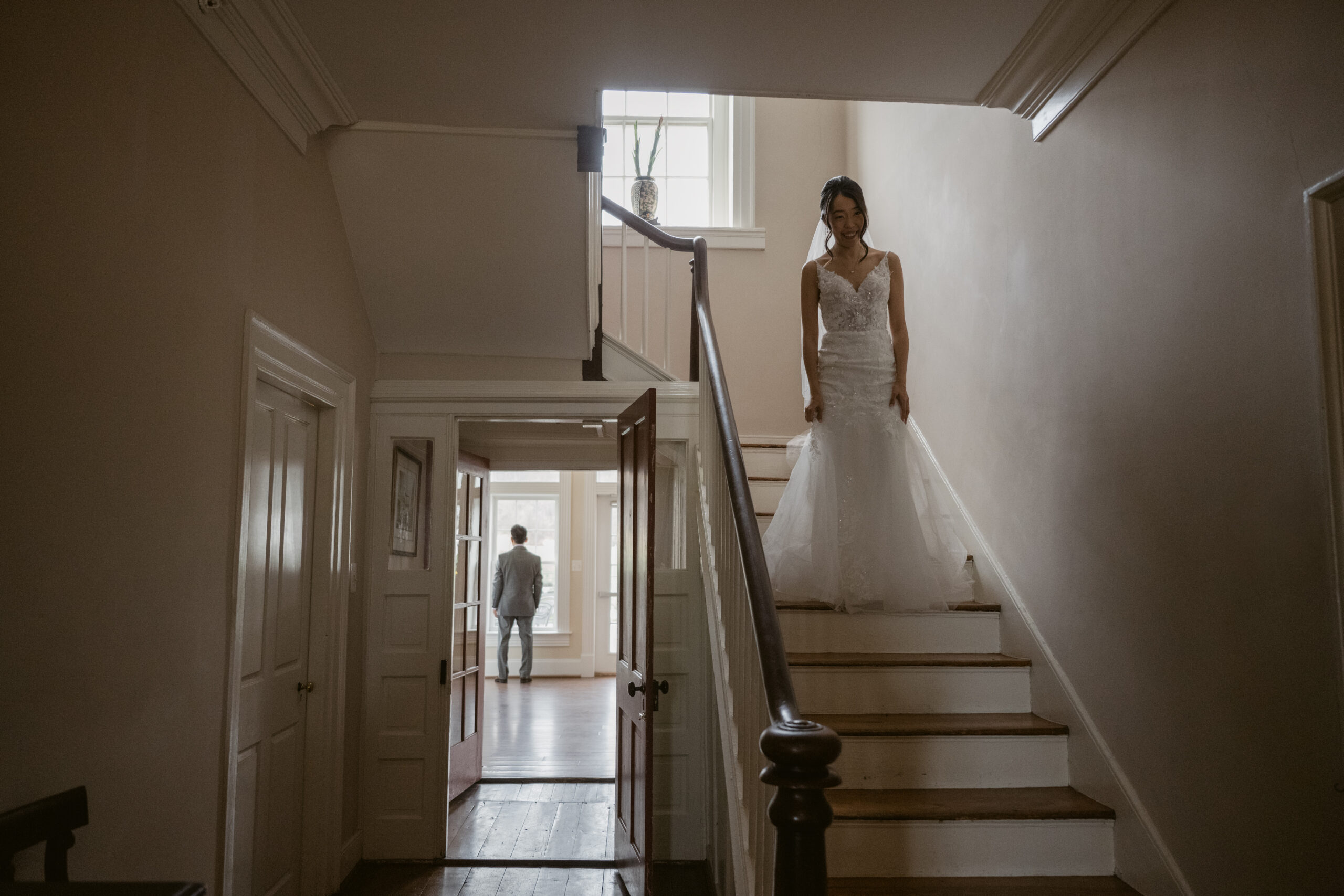 Bride descending the staircase at Springfield Manor moments before her first look with her father, who waits down the hall with his back turned.