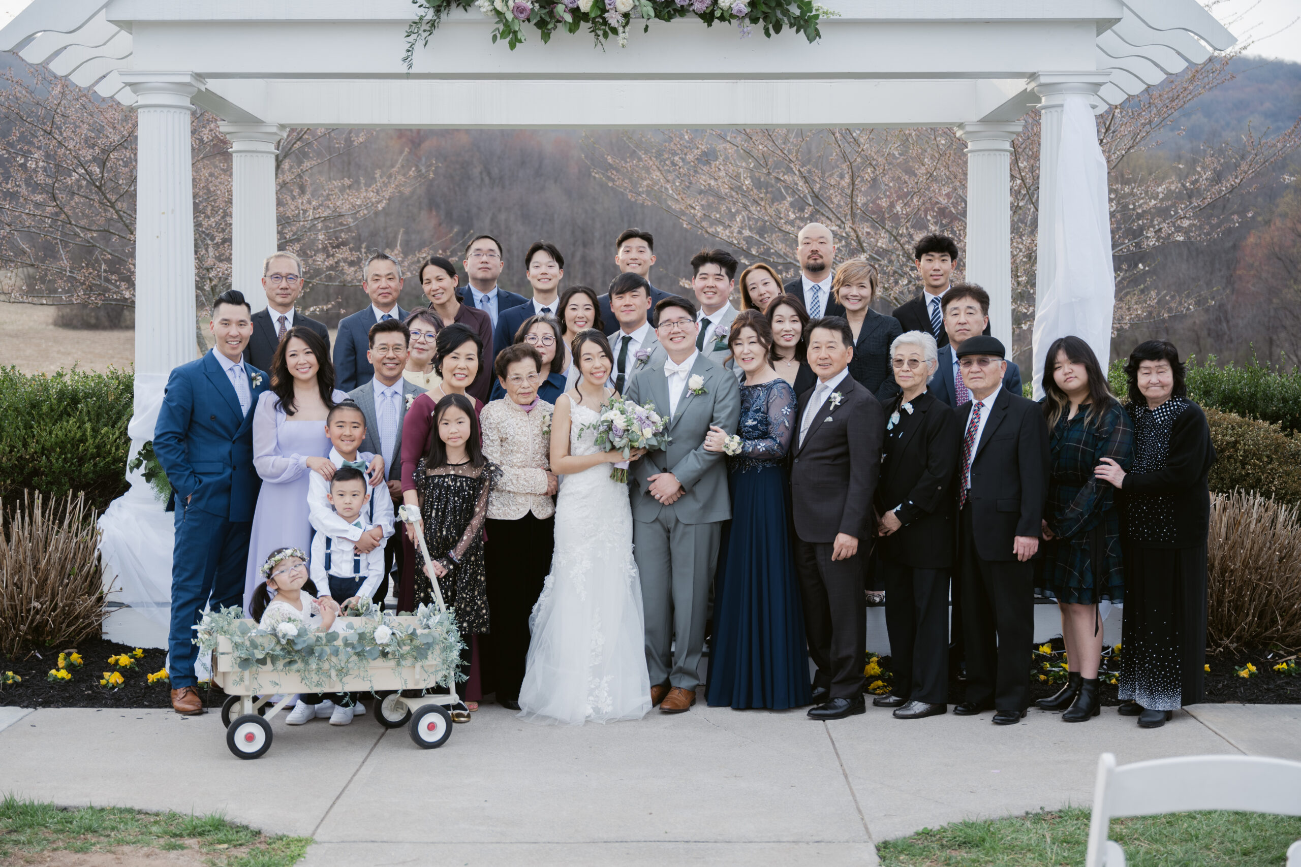 Large family wedding portrait taken under a white arbor, with the bride and groom centered.