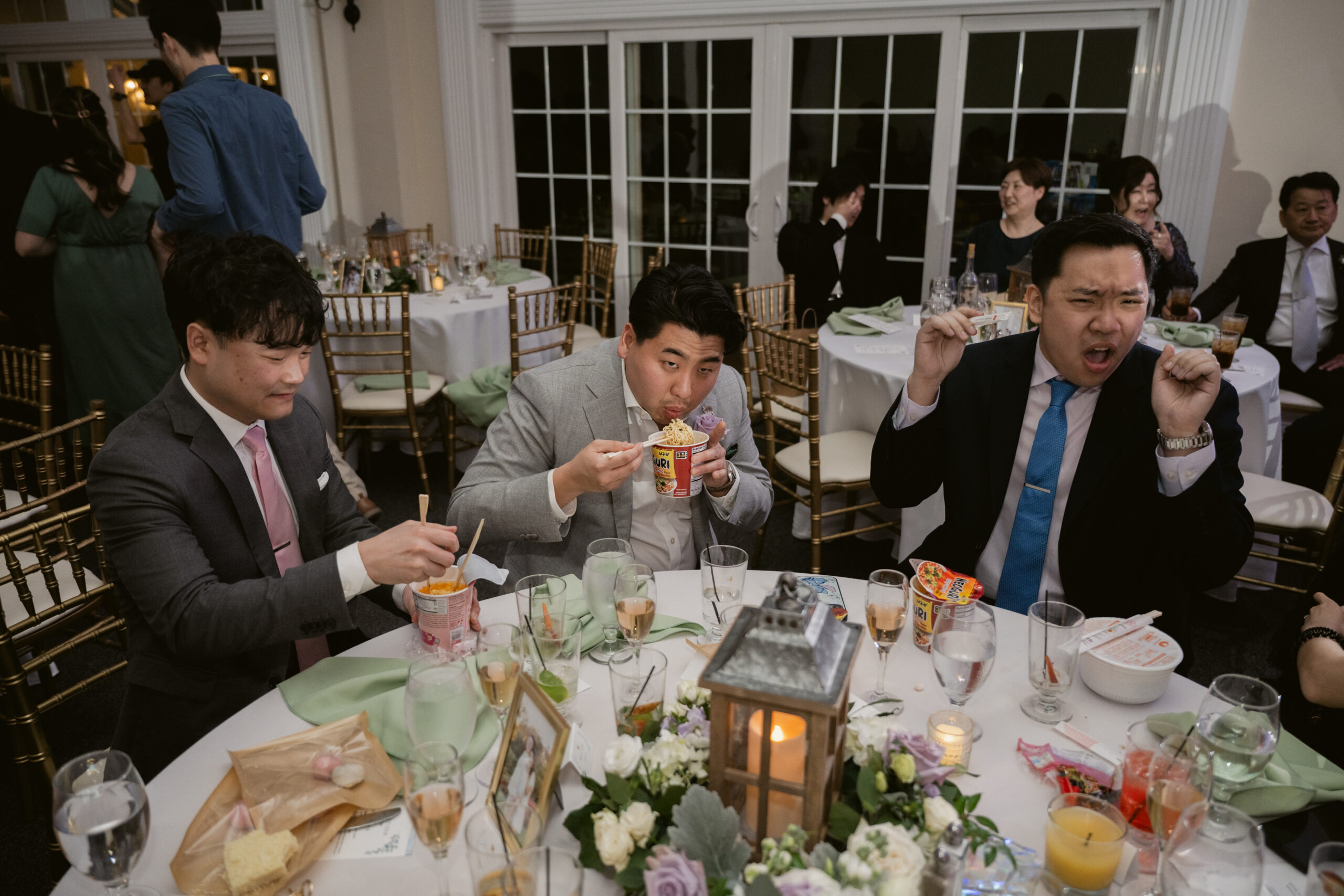 Three male wedding guests sitting at a reception table, enjoying cups of instant noodles and laughing.