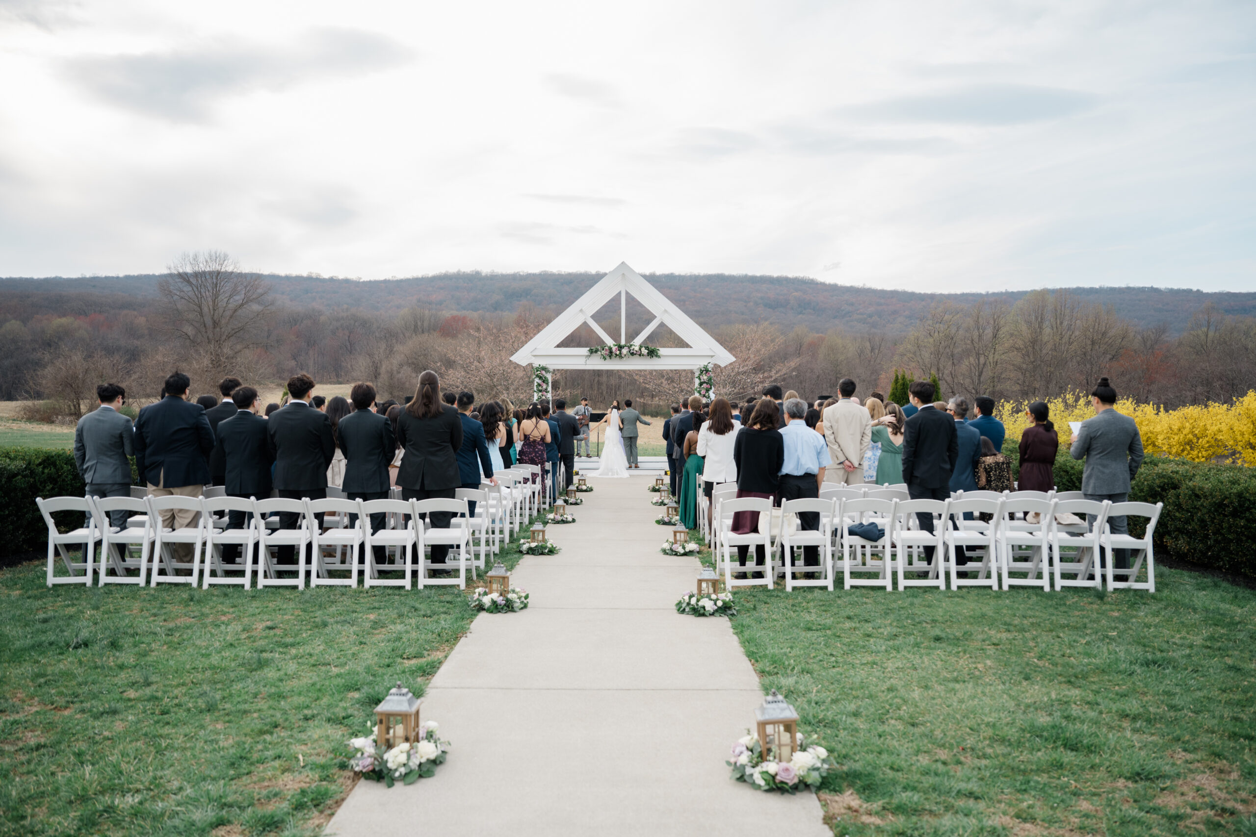 Wedding guests stand as the bride walks down the aisle toward a floral-decorated white arbor at an outdoor ceremony, framed by spring mountain views at Springfield Manor.