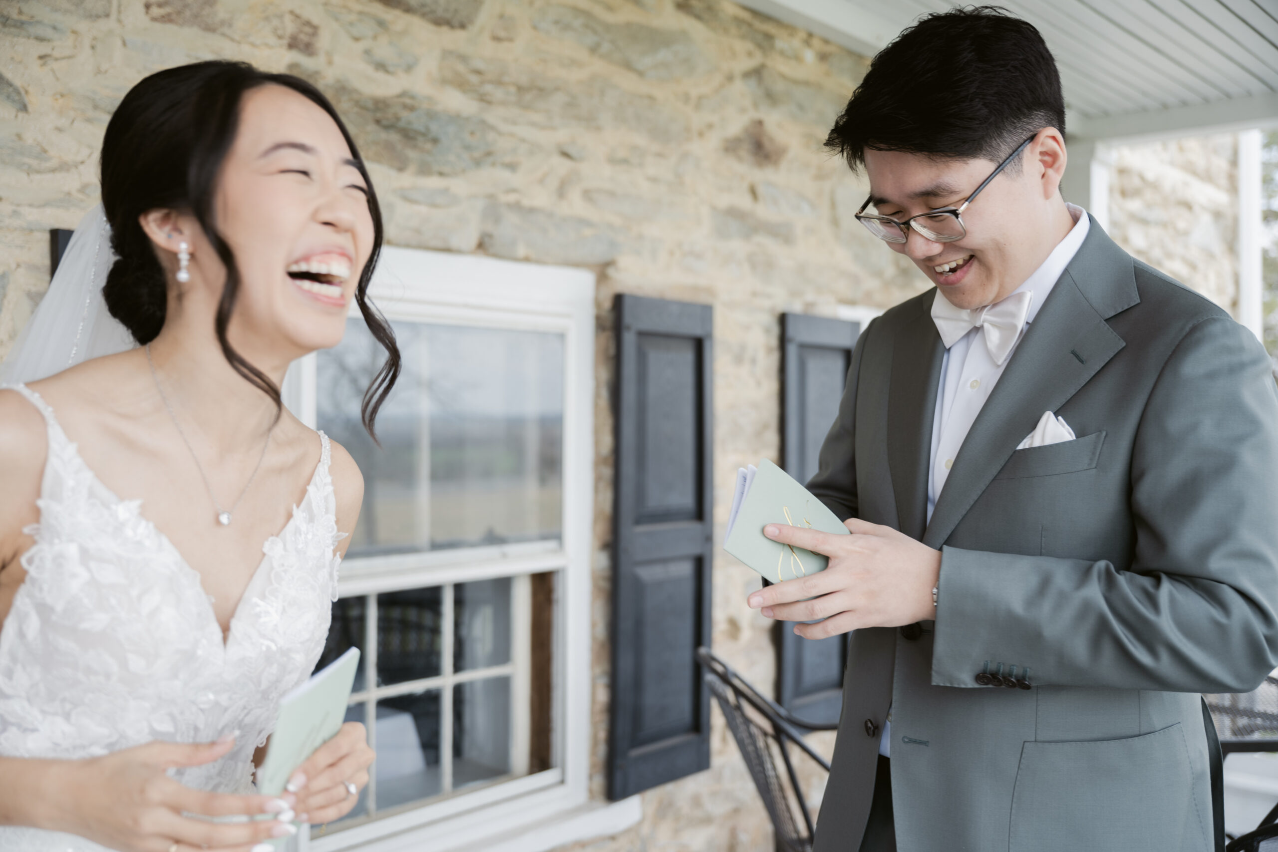Bride and groom laughing joyfully while exchanging handwritten vows during their first look outside a stone building at Springfield Manor.
