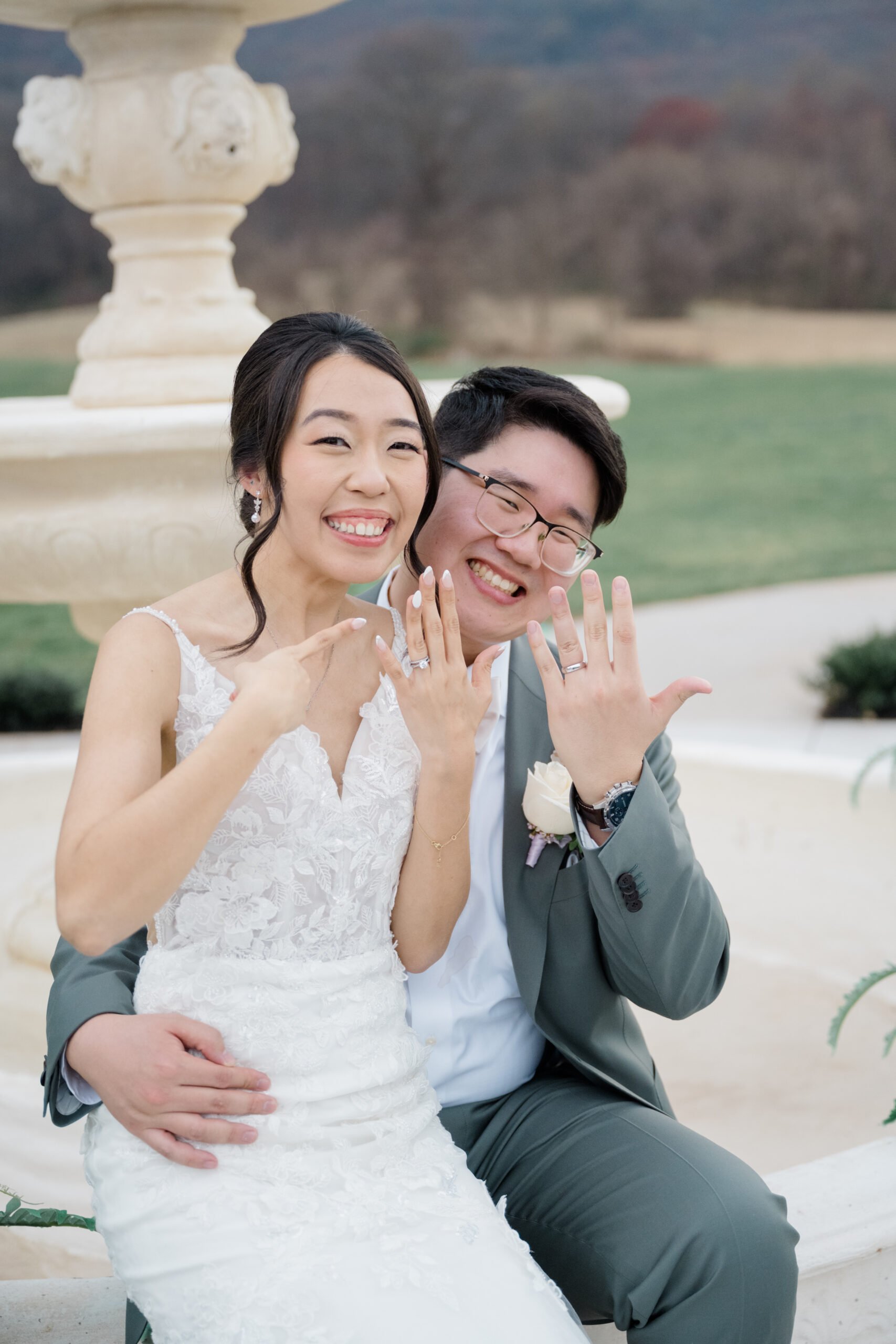 Bride and groom sitting by a fountain, smiling and pointing to their wedding rings.